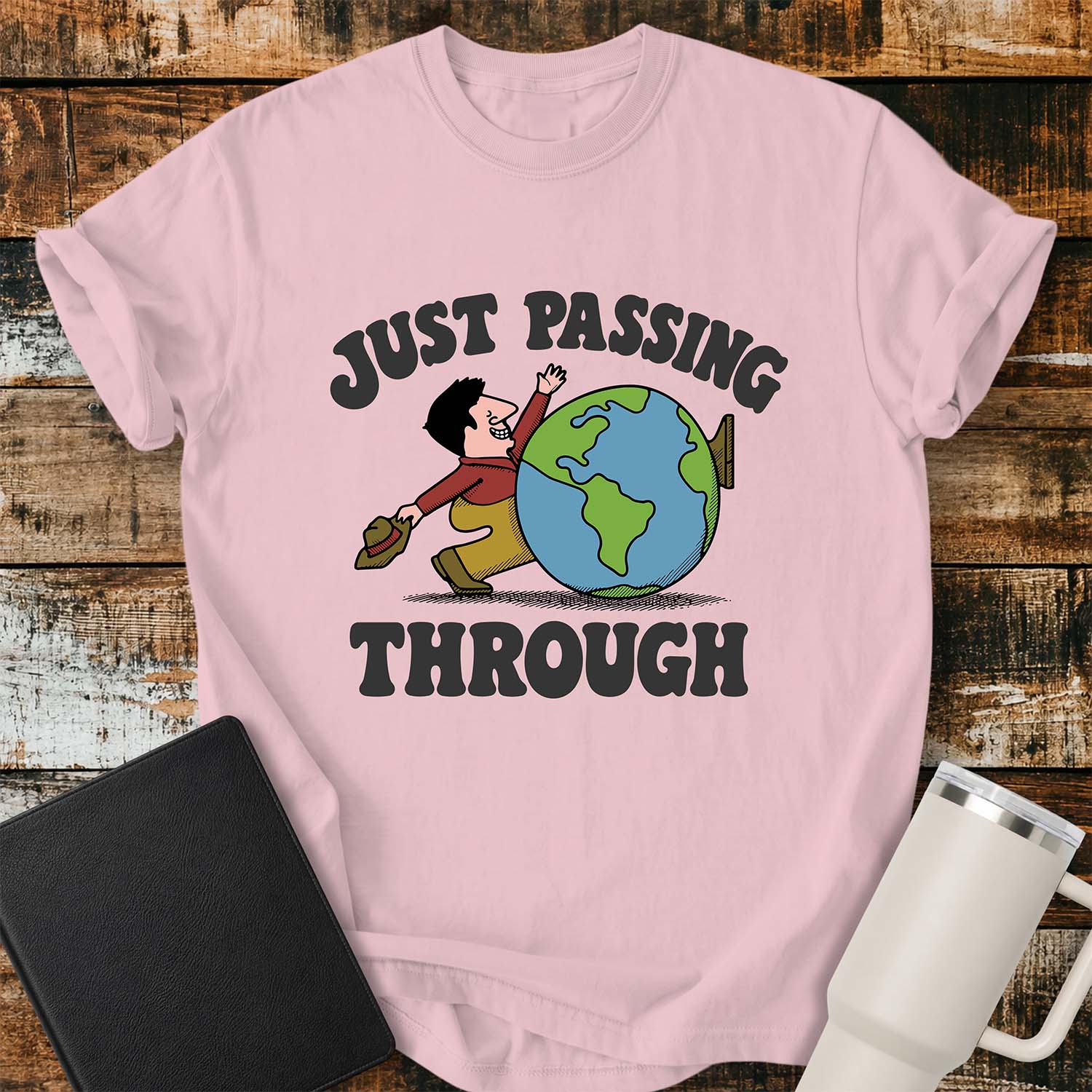 blossom pink T-shirt featuring a retro cartoon of a smiling traveler carrying a hat while walking behind a globe with the phrase “JUST PASSING THROUGH” in bold vintage lettering. The shirt is displayed on a rustic wood plank background alongside a black Bible and a white insulated tumbler.