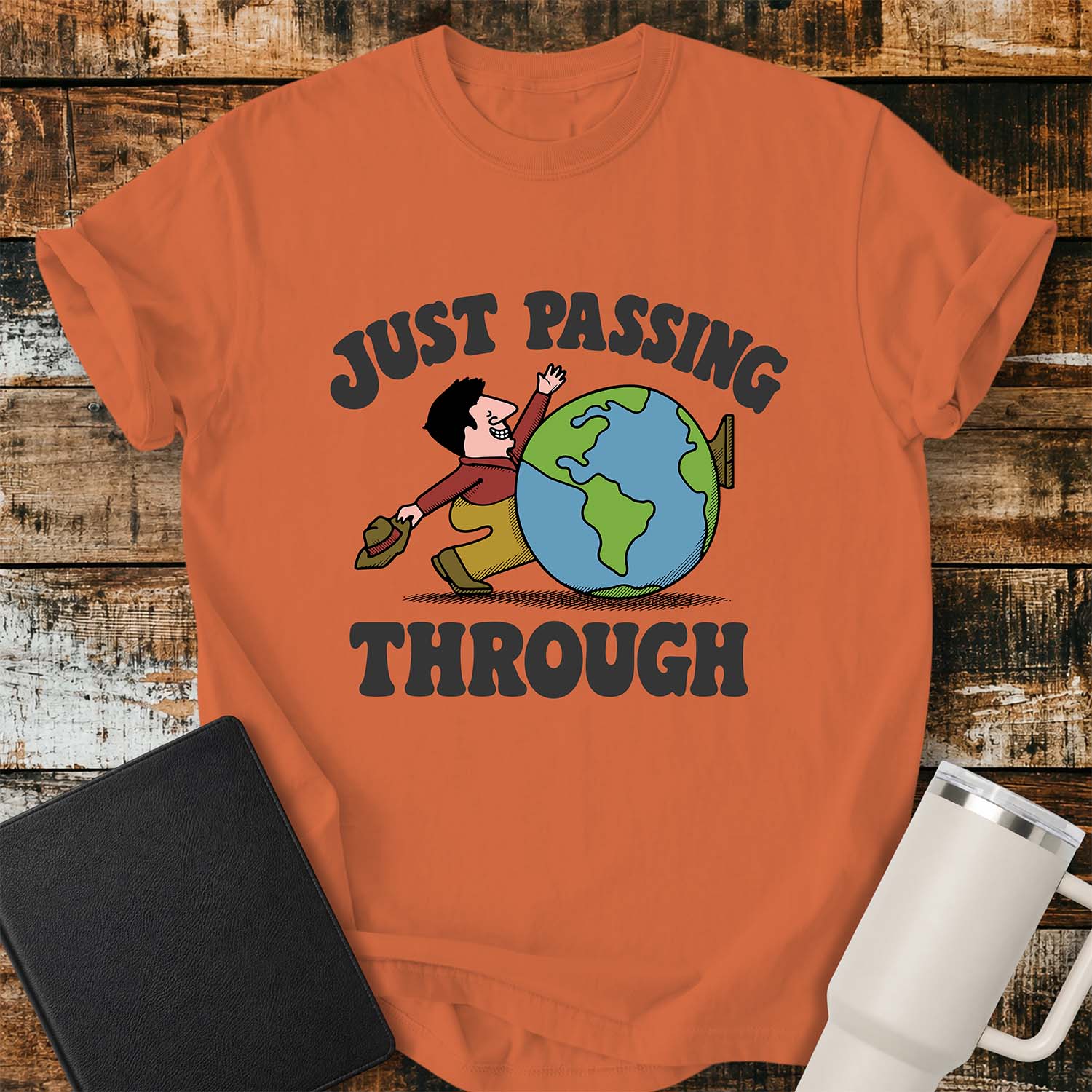 Burnt orange T-shirt featuring a retro cartoon of a smiling traveler carrying a hat while walking behind a globe with the phrase “JUST PASSING THROUGH” in bold vintage lettering. The shirt is displayed on a rustic wood plank background alongside a black Bible and a white insulated tumbler.