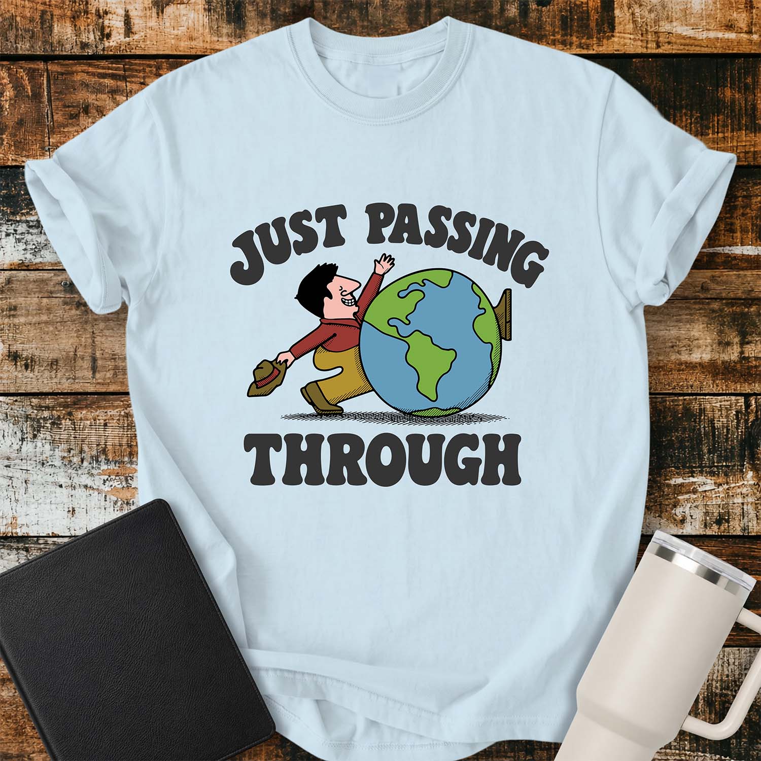 chambray blue T-shirt featuring a retro cartoon of a smiling traveler carrying a hat while walking behind a globe with the phrase “JUST PASSING THROUGH” in bold vintage lettering. The shirt is displayed on a rustic wood plank background alongside a black Bible and a white insulated tumbler.