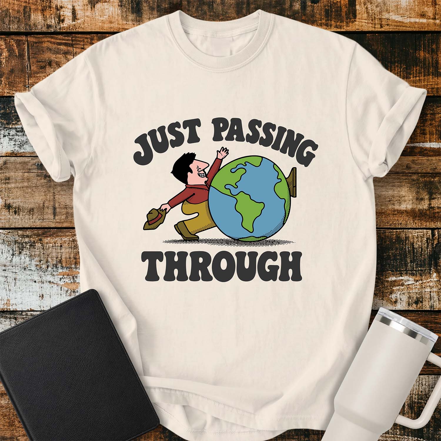 ivory T-shirt featuring a retro cartoon of a smiling traveler carrying a hat while walking behind a globe with the phrase “JUST PASSING THROUGH” in bold vintage lettering. The shirt is displayed on a rustic wood plank background alongside a black Bible and a white insulated tumbler.