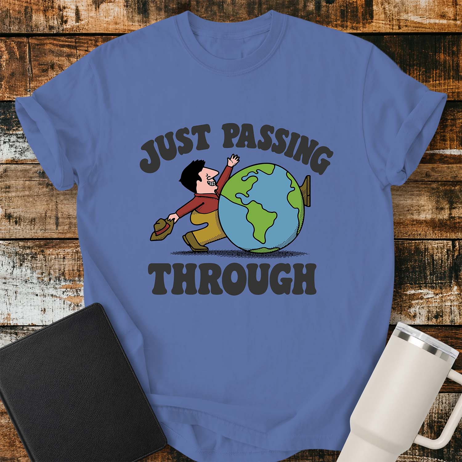 mystic blue T-shirt featuring a retro cartoon of a smiling traveler carrying a hat while walking behind a globe with the phrase “JUST PASSING THROUGH” in bold vintage lettering. The shirt is displayed on a rustic wood plank background alongside a black Bible and a white insulated tumbler.