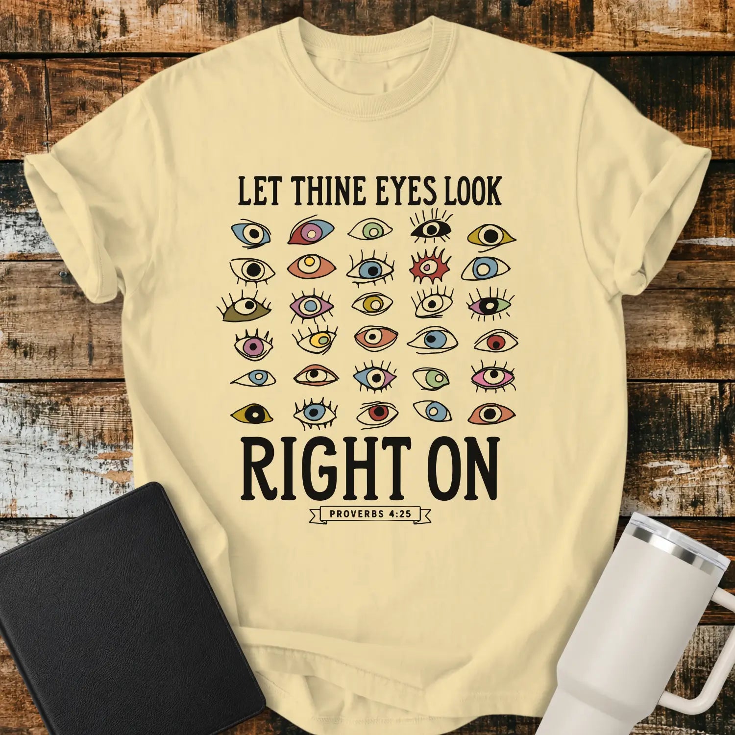 banana yellow graphic T-shirt mockup featuring the phrase “Let Thine Eyes Look Right On” with a Proverbs 4:25 reference, surrounded by a collection of colorful illustrated eyes, styled with a black Bible and white tumbler on a rustic wood background.