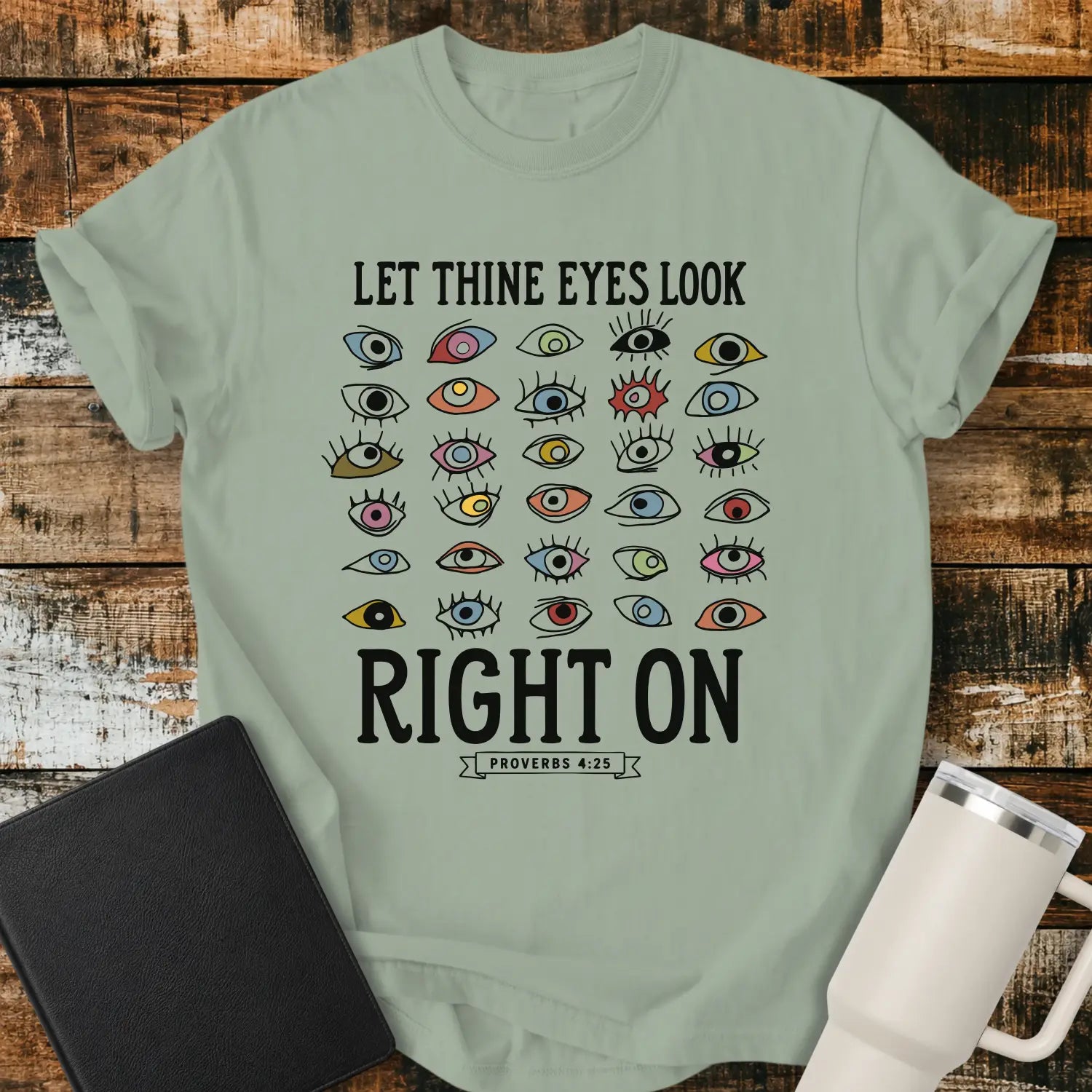 bay green graphic T-shirt mockup featuring the phrase “Let Thine Eyes Look Right On” with a Proverbs 4:25 reference, surrounded by a collection of colorful illustrated eyes, styled with a black Bible and white tumbler on a rustic wood background.
