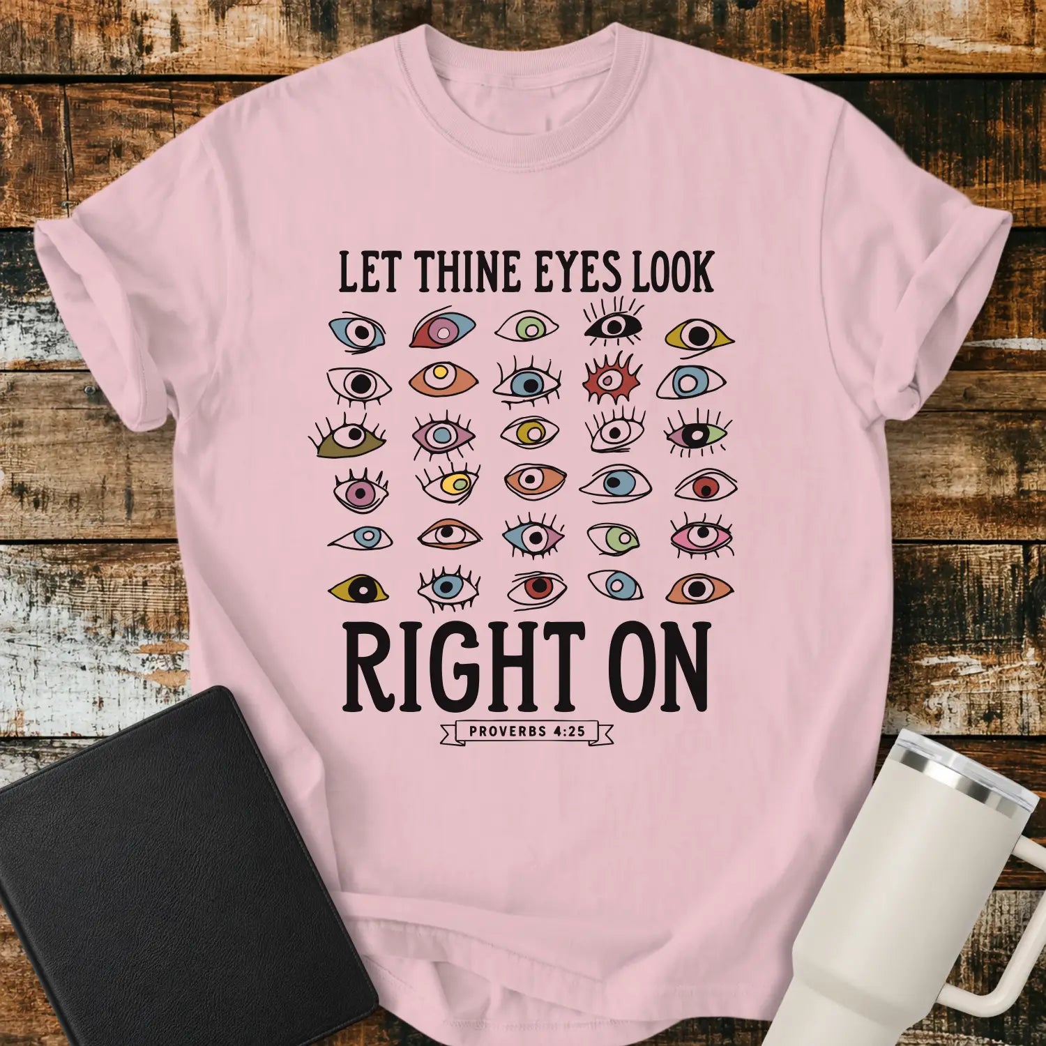 blossom pink graphic T-shirt mockup featuring the phrase “Let Thine Eyes Look Right On” with a Proverbs 4:25 reference, surrounded by a collection of colorful illustrated eyes, styled with a black Bible and white tumbler on a rustic wood background.