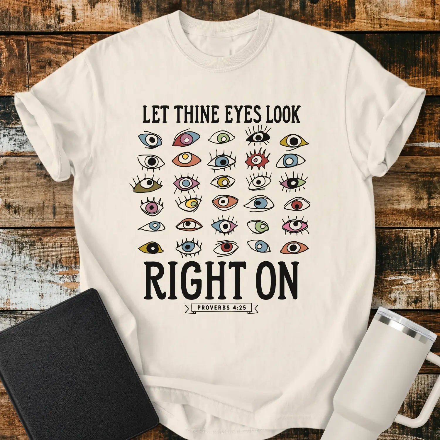 ivory graphic T-shirt mockup featuring the phrase “Let Thine Eyes Look Right On” with a Proverbs 4:25 reference, surrounded by a collection of colorful illustrated eyes, styled with a black Bible and white tumbler on a rustic wood background.