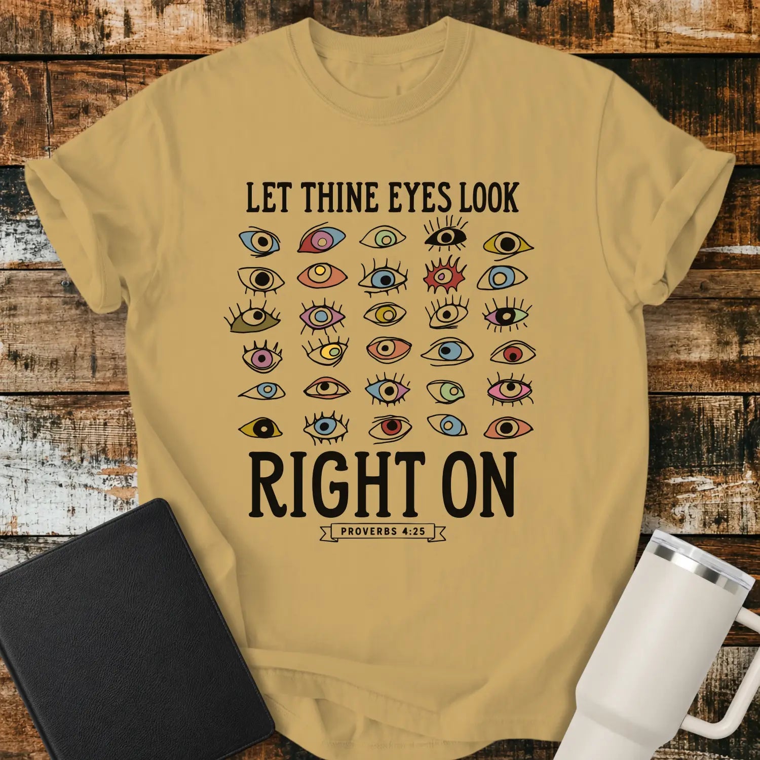 mustard graphic T-shirt mockup featuring the phrase “Let Thine Eyes Look Right On” with a Proverbs 4:25 reference, surrounded by a collection of colorful illustrated eyes, styled with a black Bible and white tumbler on a rustic wood background.