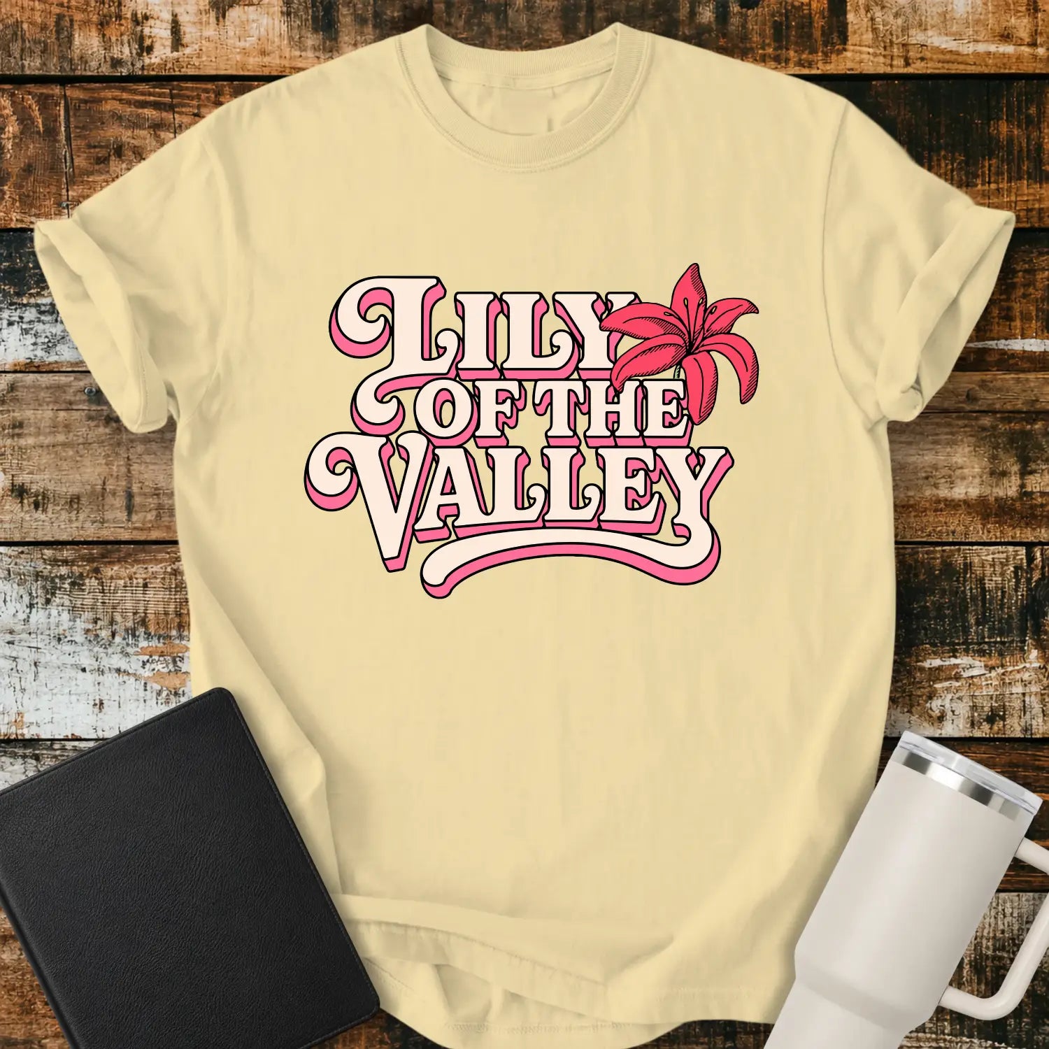 banana yellow T-shirt mockup featuring retro pink and cream typography reading “Lily of the Valley,” accented with a large pink lily flower. The shirt is displayed on a rustic wood plank background alongside a black Bible and a white insulated tumbler, creating a casual Christian apparel presentation.