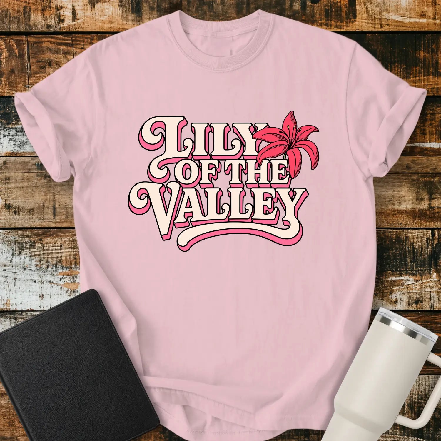 blossom pink T-shirt mockup featuring retro pink and cream typography reading “Lily of the Valley,” accented with a large pink lily flower. The shirt is displayed on a rustic wood plank background alongside a black Bible and a white insulated tumbler, creating a casual Christian apparel presentation.