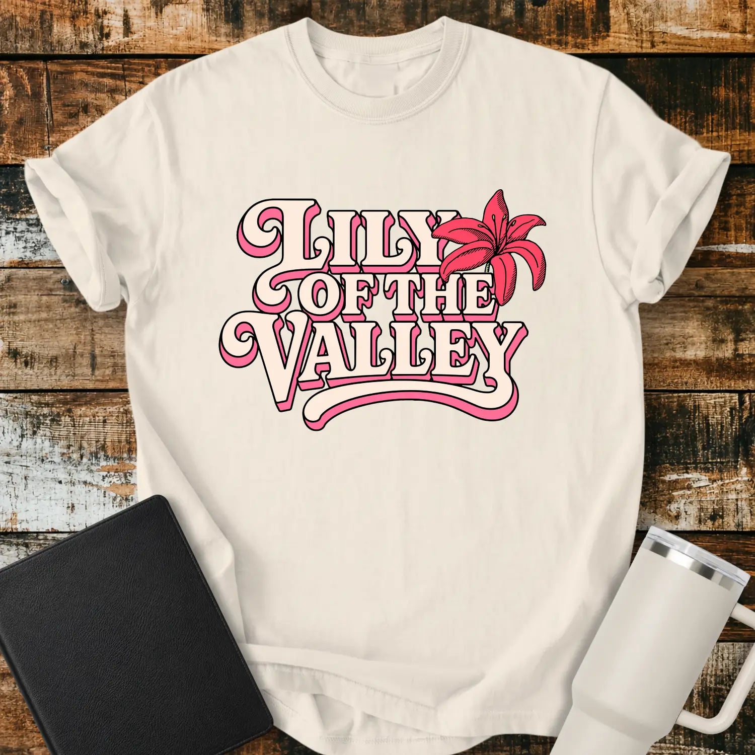 ivory T-shirt mockup featuring retro pink and cream typography reading “Lily of the Valley,” accented with a large pink lily flower. The shirt is displayed on a rustic wood plank background alongside a black Bible and a white insulated tumbler, creating a casual Christian apparel presentation.