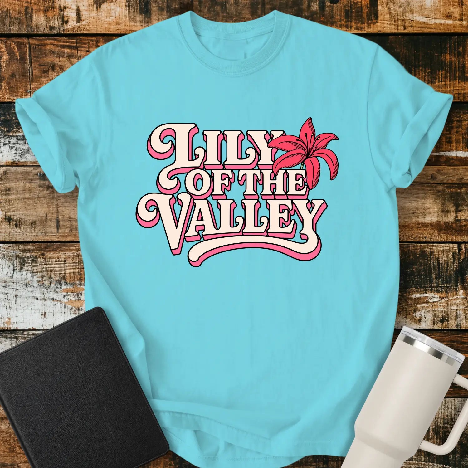 Lagoon blue T-shirt mockup featuring retro pink and cream typography reading “Lily of the Valley,” accented with a large pink lily flower. The shirt is displayed on a rustic wood plank background alongside a black Bible and a white insulated tumbler, creating a casual Christian apparel presentation.