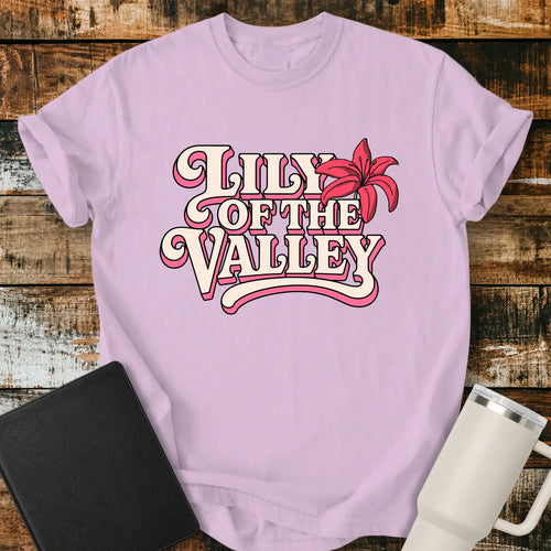 orchid purple T-shirt mockup featuring retro pink and cream typography reading “Lily of the Valley,” accented with a large pink lily flower. The shirt is displayed on a rustic wood plank background alongside a black Bible and a white insulated tumbler, creating a casual Christian apparel presentation.