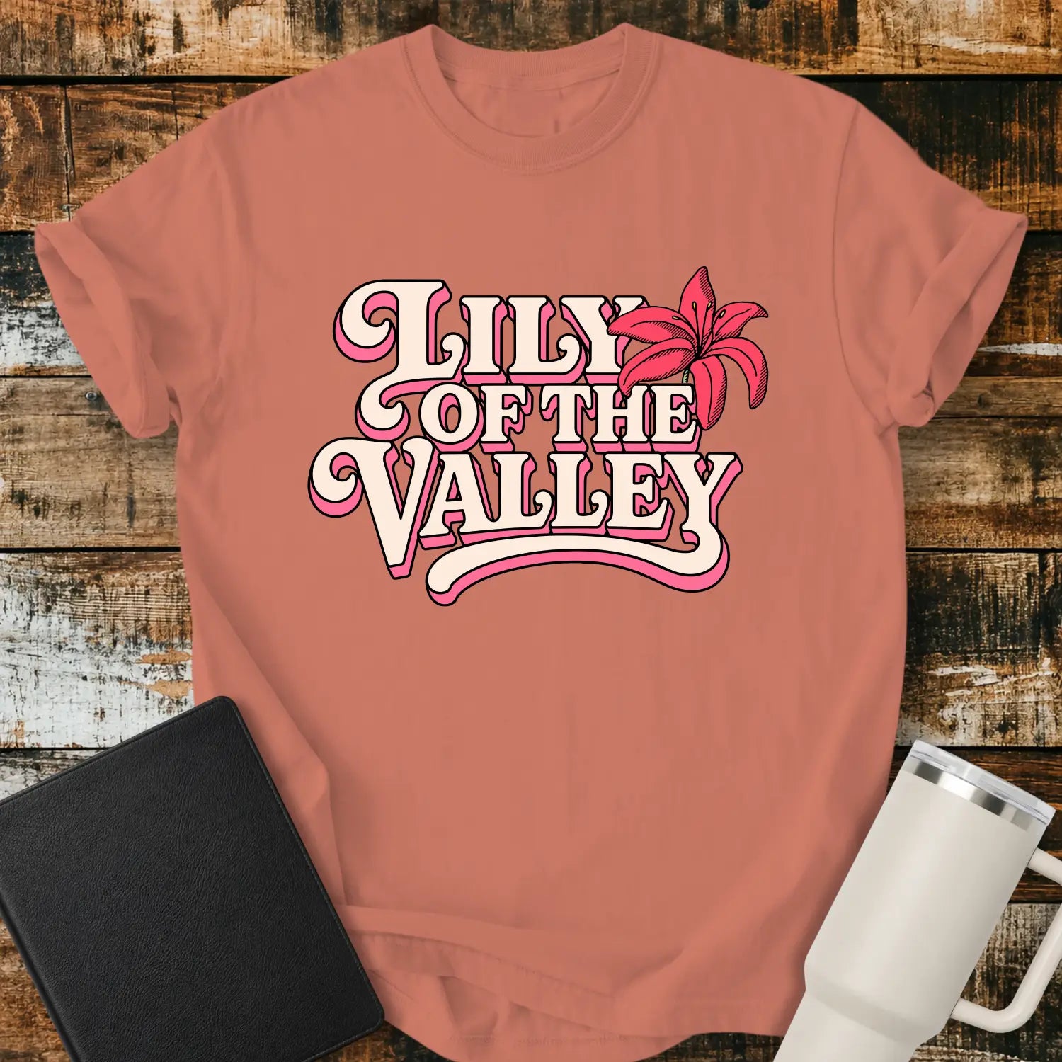 terracotta T-shirt mockup featuring retro pink and cream typography reading “Lily of the Valley,” accented with a large pink lily flower. The shirt is displayed on a rustic wood plank background alongside a black Bible and a white insulated tumbler, creating a casual Christian apparel presentation.