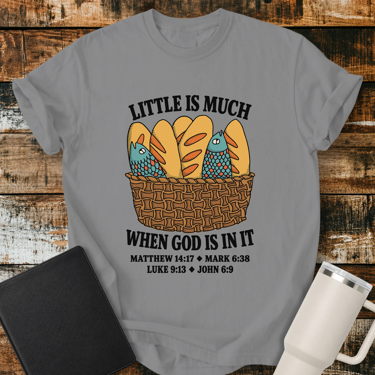 grey Christian t-shirt featuring a vintage-style basket filled with five loaves and two fish, with bold lettering reading “Little Is Much When God Is in It” and references to Matthew 14:17, Mark 6:38, Luke 9:13, and John 6:9.