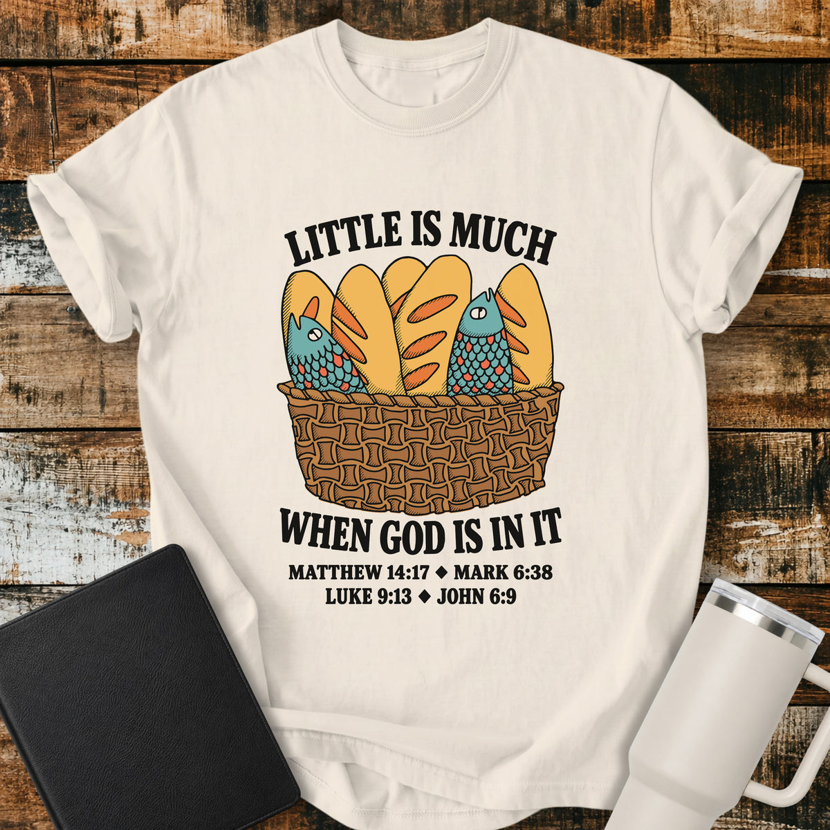 Ivory Christian t-shirt featuring a vintage-style basket filled with five loaves and two fish, with bold lettering reading “Little Is Much When God Is in It” and references to Matthew 14:17, Mark 6:38, Luke 9:13, and John 6:9.