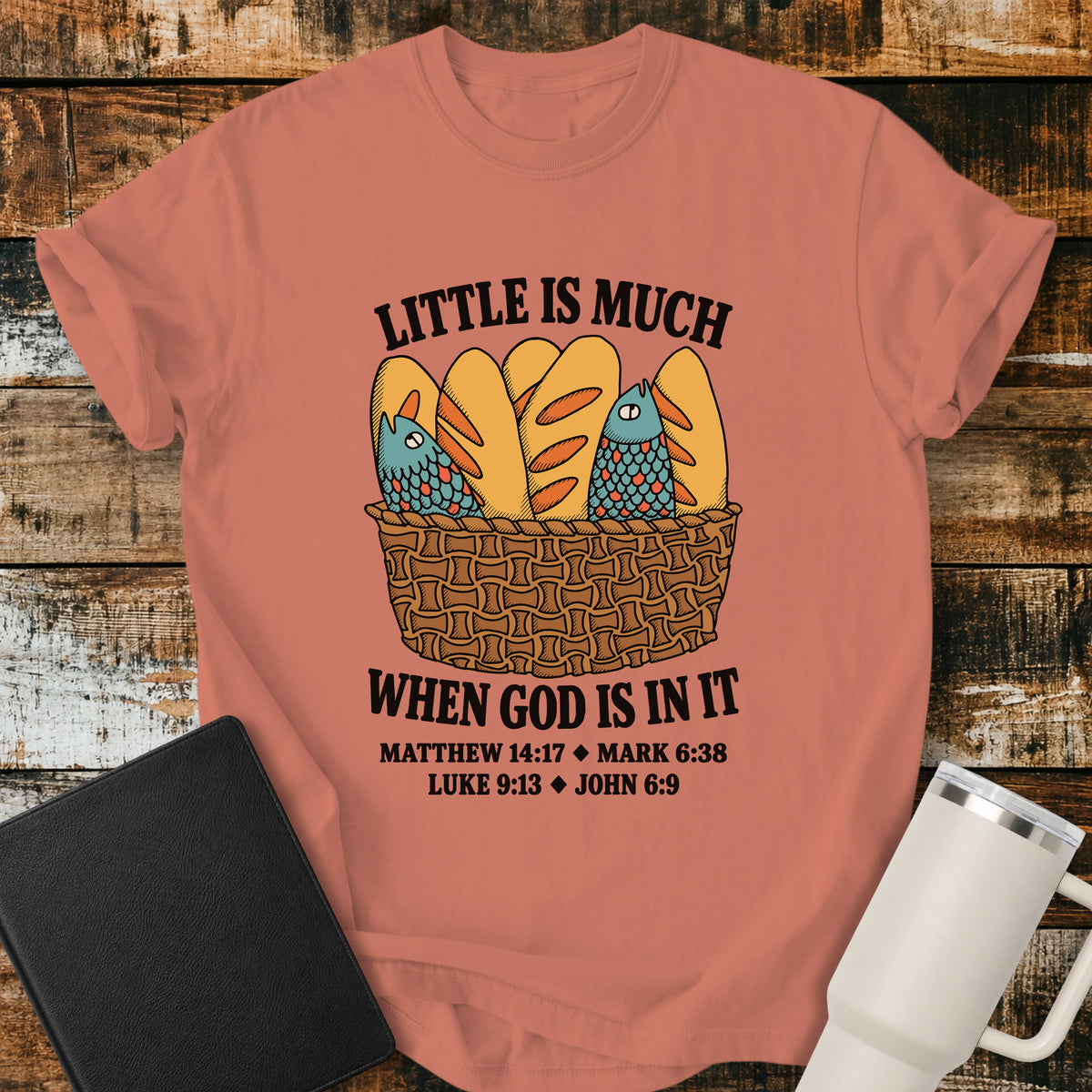 treracotta Christian t-shirt featuring a vintage-style basket filled with five loaves and two fish, with bold lettering reading “Little Is Much When God Is in It” and references to Matthew 14:17, Mark 6:38, Luke 9:13, and John 6:9.