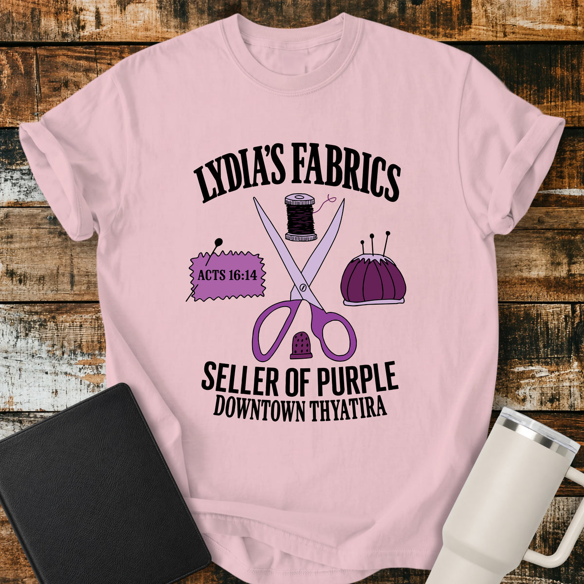 blossom pink Christian t-shirt featuring vintage sewing notions—scissors, thread, pincushion, thimble, and fabric—with retro shop-style lettering reading “Lydia’s Fabrics,” “Seller of Purple,” “Downtown Thyatira,” and Acts 16:14.