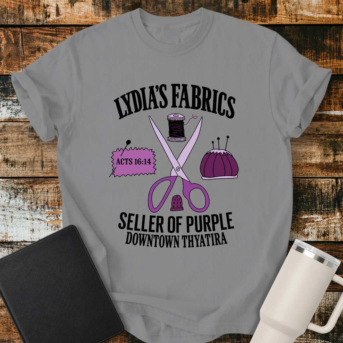 grey Christian t-shirt featuring vintage sewing notions—scissors, thread, pincushion, thimble, and fabric—with retro shop-style lettering reading “Lydia’s Fabrics,” “Seller of Purple,” “Downtown Thyatira,” and Acts 16:14.