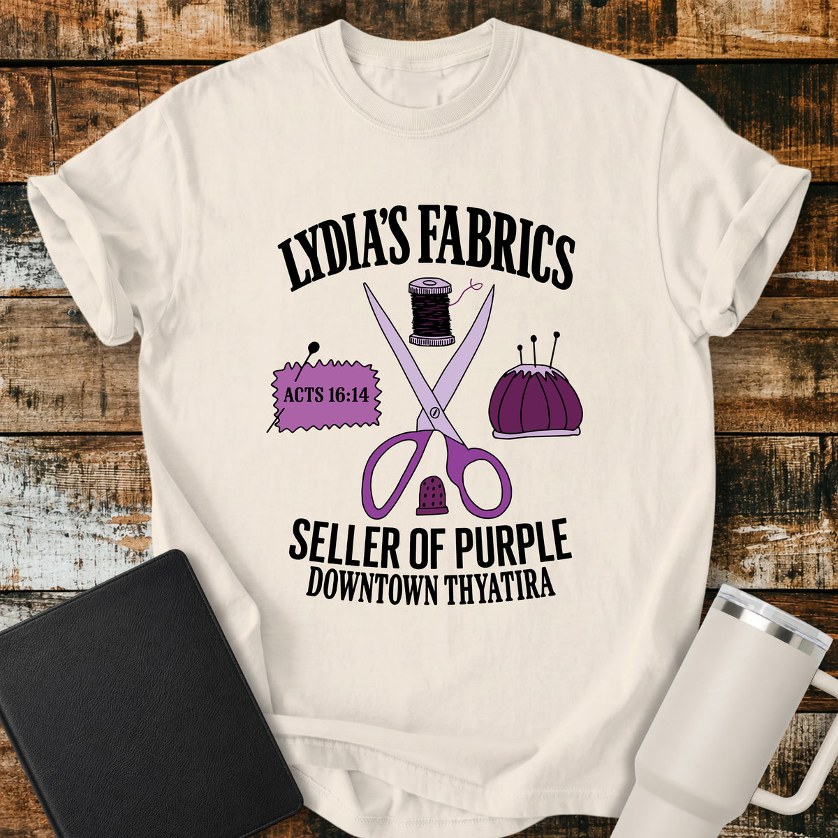 ivory Christian t-shirt featuring vintage sewing notions—scissors, thread, pincushion, thimble, and fabric—with retro shop-style lettering reading “Lydia’s Fabrics,” “Seller of Purple,” “Downtown Thyatira,” and Acts 16:14.