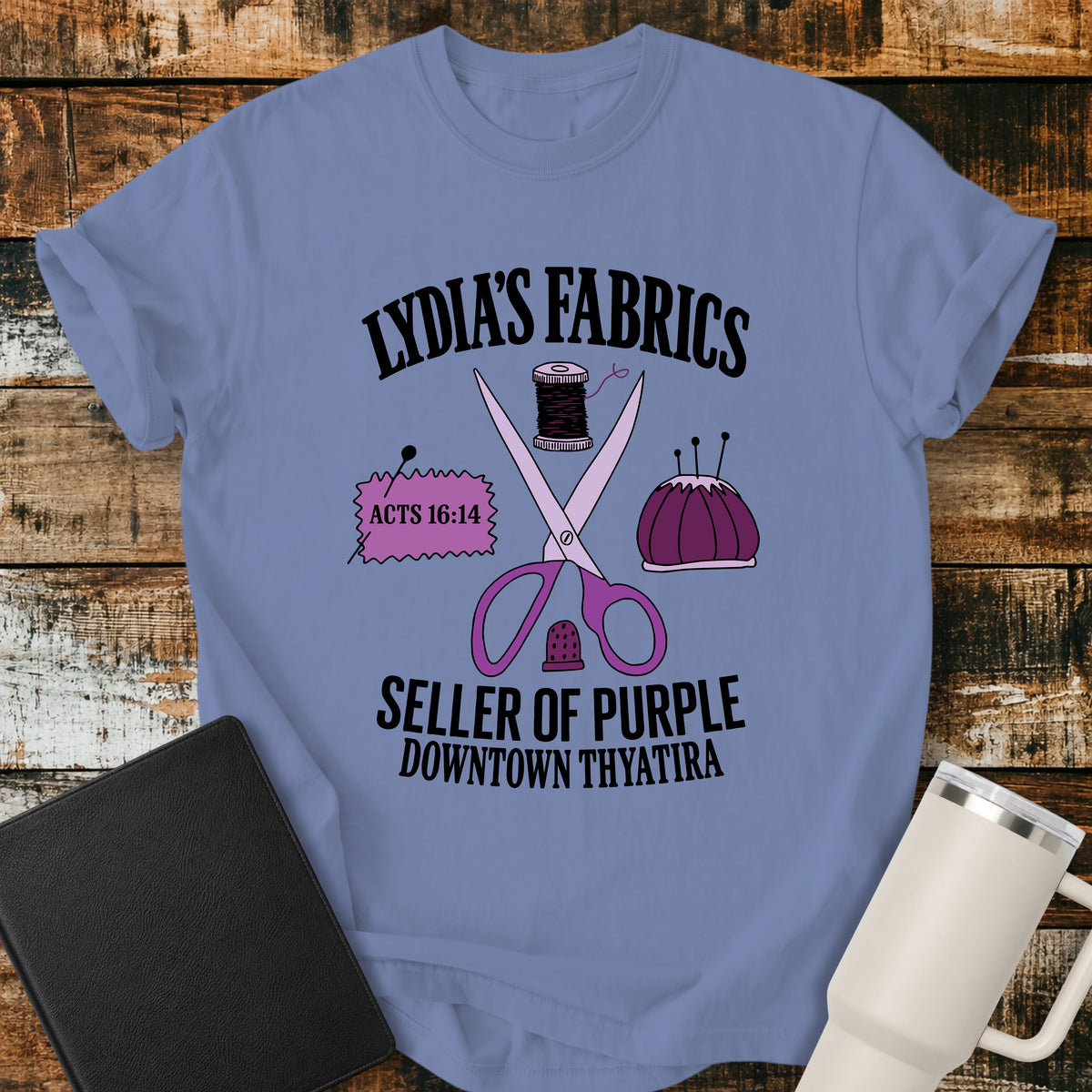 washed denim blue Christian t-shirt featuring vintage sewing notions—scissors, thread, pincushion, thimble, and fabric—with retro shop-style lettering reading “Lydia’s Fabrics,” “Seller of Purple,” “Downtown Thyatira,” and Acts 16:14.