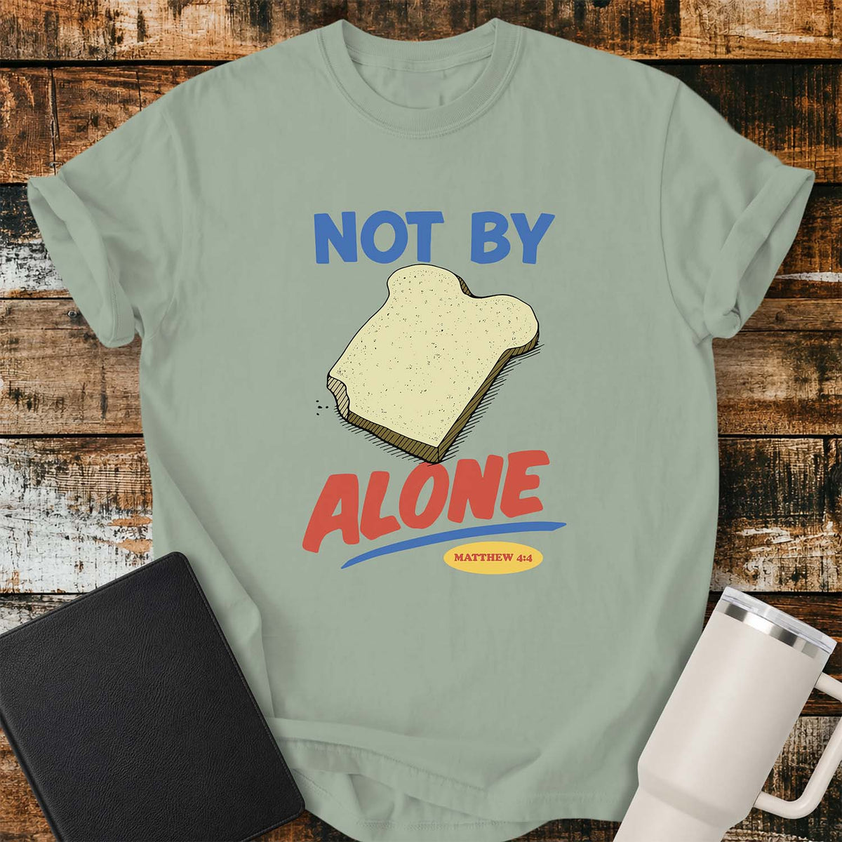 bay green T-shirt featuring a colorful illustration of a slice of bread between the words “NOT BY” and “ALONE,” referencing Matthew 4:4. The phrase “MATTHEW 4:4” appears in a small yellow oval beneath the word “ALONE.” The design uses retro blue and orange lettering to emphasize the biblical message that man does not live by bread alone. The shirt is displayed on a rustic wood plank background with a black Bible on the left and a white insulated tumbler on the right.