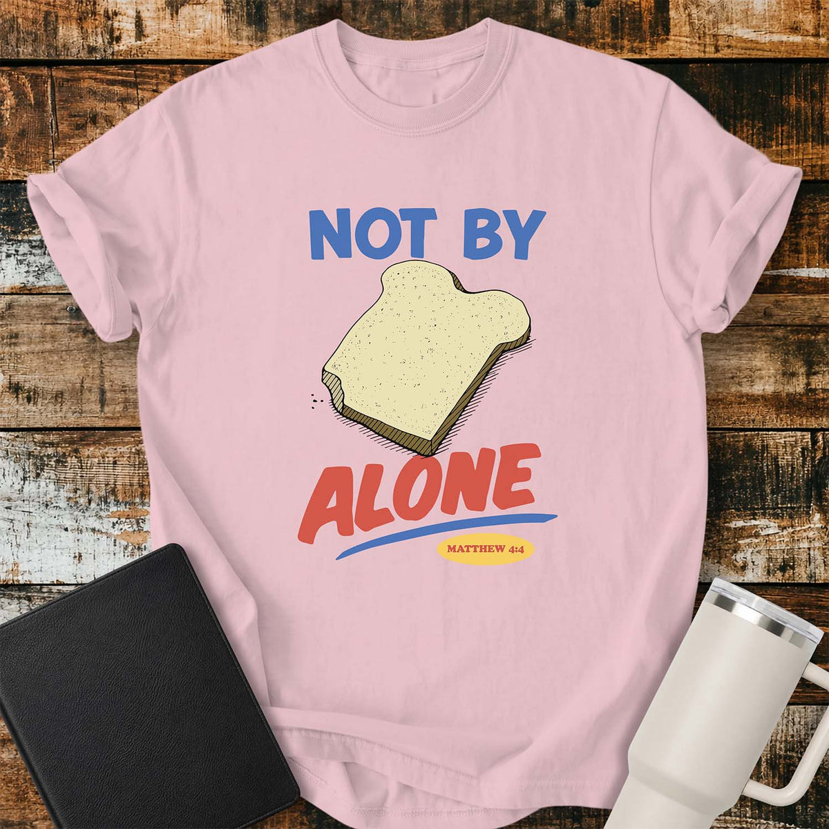 blossom pink T-shirt featuring a colorful illustration of a slice of bread between the words “NOT BY” and “ALONE,” referencing Matthew 4:4. The phrase “MATTHEW 4:4” appears in a small yellow oval beneath the word “ALONE.” The design uses retro blue and orange lettering to emphasize the biblical message that man does not live by bread alone. The shirt is displayed on a rustic wood plank background with a black Bible on the left and a white insulated tumbler on the right.