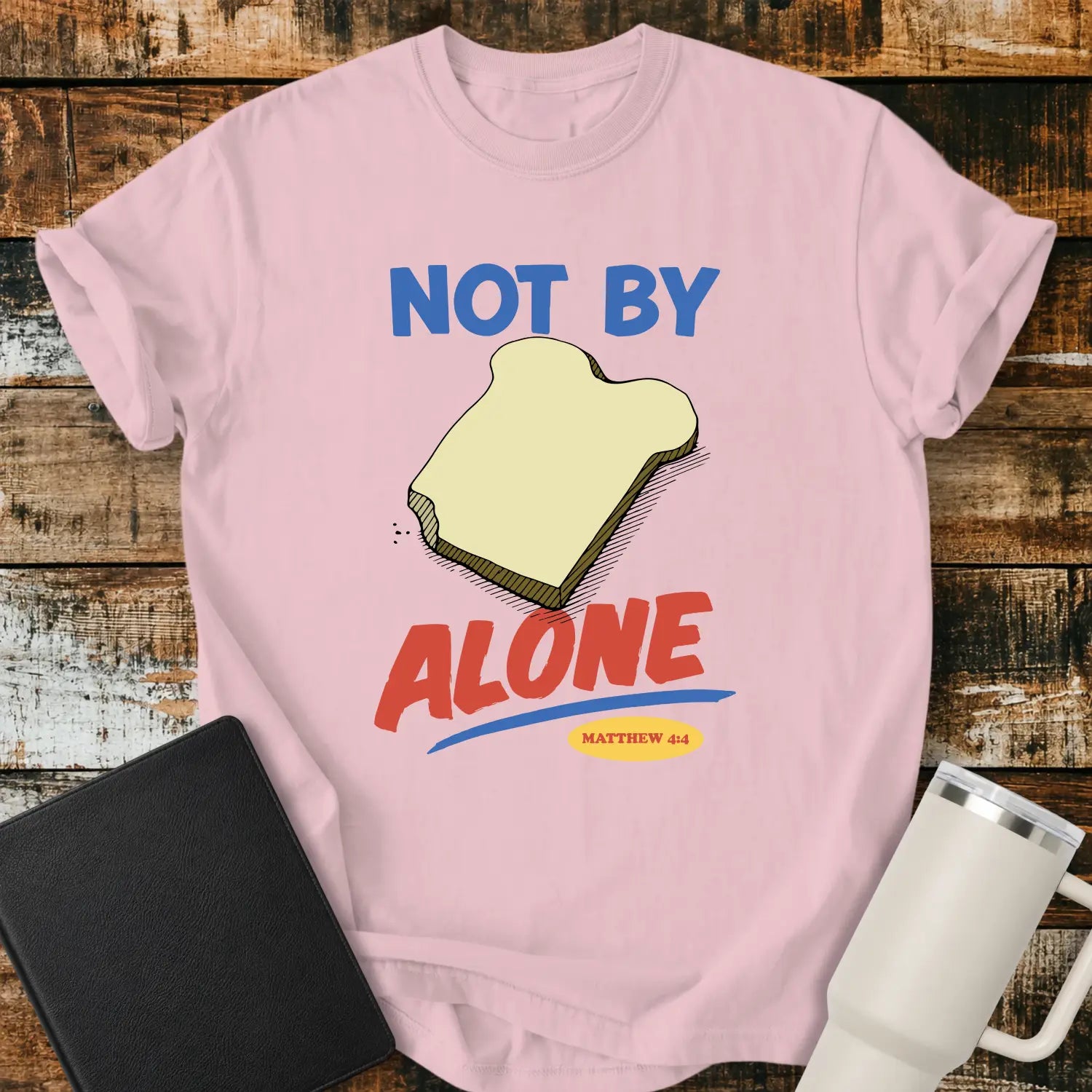 blossom pink T-shirt mockup featuring the phrase “NOT BY BREAD ALONE” inspired by Matthew 4:4. The design includes a slice of bread with simple hand-drawn shading, blue and red retro lettering, and a small Matthew 4:4 reference. The shirt is displayed on a rustic wood plank background beside a black Bible and a white insulated tumbler.
