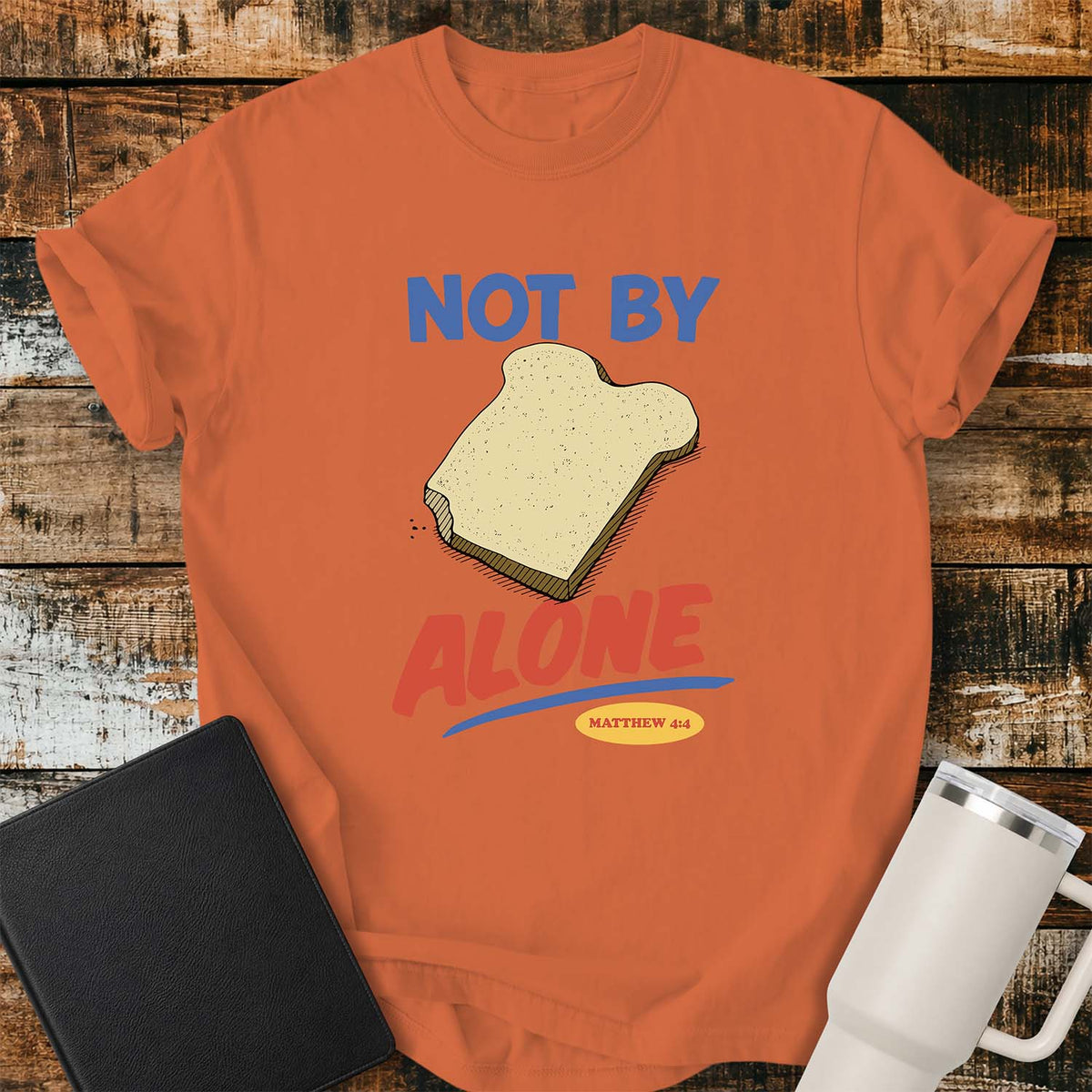 burnt orange T-shirt featuring a colorful illustration of a slice of bread between the words “NOT BY” and “ALONE,” referencing Matthew 4:4. The phrase “MATTHEW 4:4” appears in a small yellow oval beneath the word “ALONE.” The design uses retro blue and orange lettering to emphasize the biblical message that man does not live by bread alone. The shirt is displayed on a rustic wood plank background with a black Bible on the left and a white insulated tumbler on the right.
