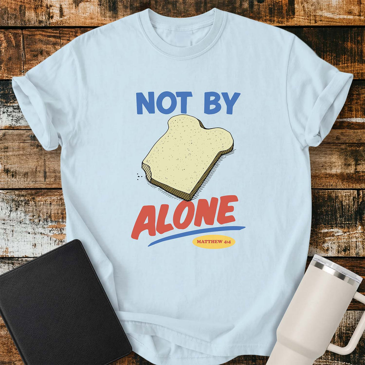 Chambray blue T-shirt featuring a colorful illustration of a slice of bread between the words “NOT BY” and “ALONE,” referencing Matthew 4:4. The phrase “MATTHEW 4:4” appears in a small yellow oval beneath the word “ALONE.” The design uses retro blue and orange lettering to emphasize the biblical message that man does not live by bread alone. The shirt is displayed on a rustic wood plank background with a black Bible on the left and a white insulated tumbler on the right.