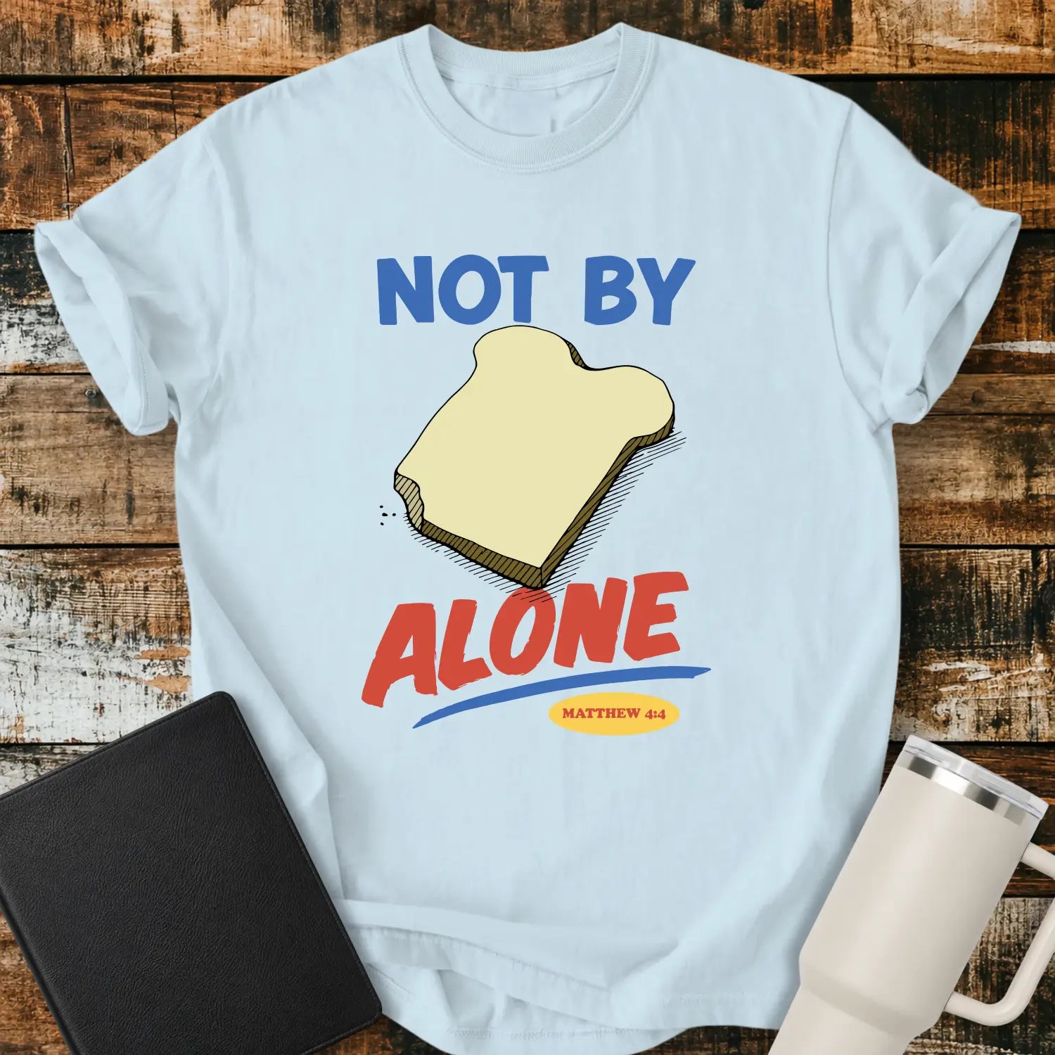 chambray blue T-shirt mockup featuring the phrase “NOT BY BREAD ALONE” inspired by Matthew 4:4. The design includes a slice of bread with simple hand-drawn shading, blue and red retro lettering, and a small Matthew 4:4 reference. The shirt is displayed on a rustic wood plank background beside a black Bible and a white insulated tumbler.
