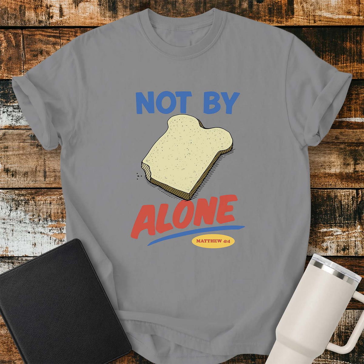 grey T-shirt featuring a colorful illustration of a slice of bread between the words “NOT BY” and “ALONE,” referencing Matthew 4:4. The phrase “MATTHEW 4:4” appears in a small yellow oval beneath the word “ALONE.” The design uses retro blue and orange lettering to emphasize the biblical message that man does not live by bread alone. The shirt is displayed on a rustic wood plank background with a black Bible on the left and a white insulated tumbler on the right.