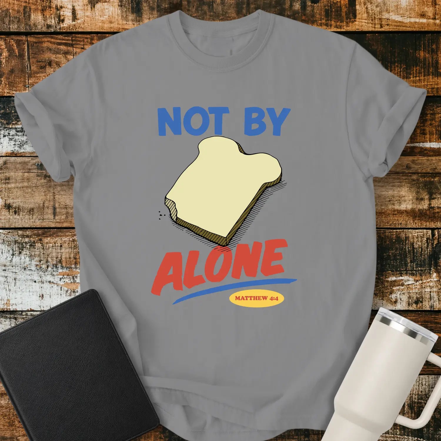 grey T-shirt mockup featuring the phrase “NOT BY BREAD ALONE” inspired by Matthew 4:4. The design includes a slice of bread with simple hand-drawn shading, blue and red retro lettering, and a small Matthew 4:4 reference. The shirt is displayed on a rustic wood plank background beside a black Bible and a white insulated tumbler.
