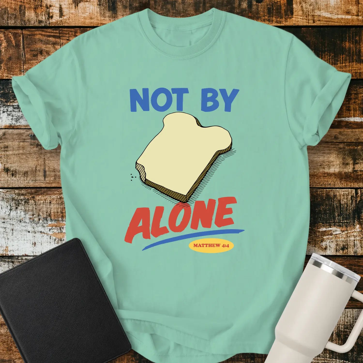 island reef green T-shirt mockup featuring the phrase “NOT BY BREAD ALONE” inspired by Matthew 4:4. The design includes a slice of bread with simple hand-drawn shading, blue and red retro lettering, and a small Matthew 4:4 reference. The shirt is displayed on a rustic wood plank background beside a black Bible and a white insulated tumbler.
