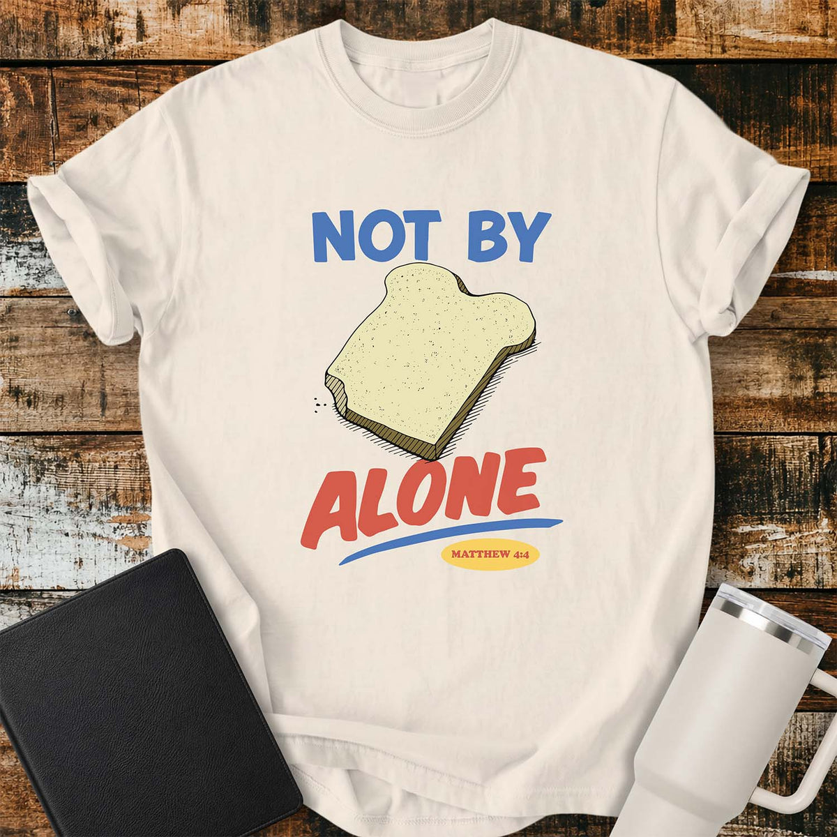 ivory T-shirt featuring a colorful illustration of a slice of bread between the words “NOT BY” and “ALONE,” referencing Matthew 4:4. The phrase “MATTHEW 4:4” appears in a small yellow oval beneath the word “ALONE.” The design uses retro blue and orange lettering to emphasize the biblical message that man does not live by bread alone. The shirt is displayed on a rustic wood plank background with a black Bible on the left and a white insulated tumbler on the right