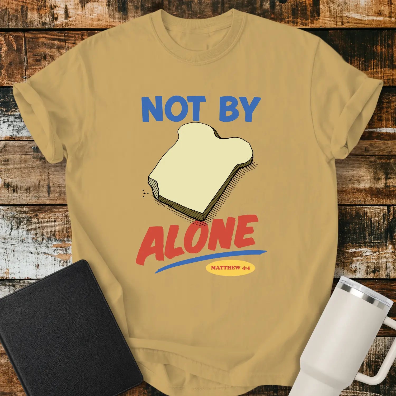 Mustard yellow T-shirt mockup featuring the phrase “NOT BY BREAD ALONE” inspired by Matthew 4:4. The design includes a slice of bread with simple hand-drawn shading, blue and red retro lettering, and a small Matthew 4:4 reference. The shirt is displayed on a rustic wood plank background beside a black Bible and a white insulated tumbler.
