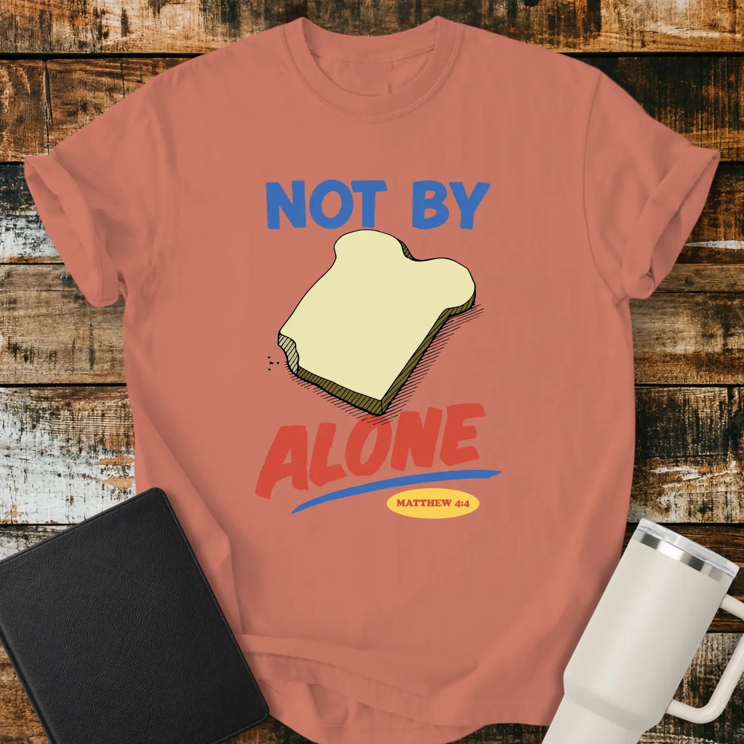 terracotta T-shirt mockup featuring the phrase “NOT BY BREAD ALONE” inspired by Matthew 4:4. The design includes a slice of bread with simple hand-drawn shading, blue and red retro lettering, and a small Matthew 4:4 reference. The shirt is displayed on a rustic wood plank background beside a black Bible and a white insulated tumbler.
