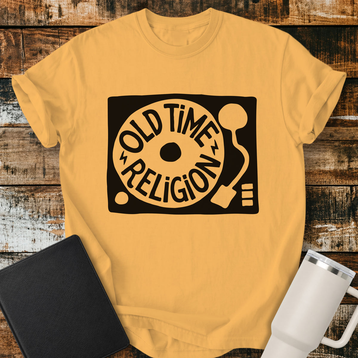 citrus yellow flat-lay t-shirt mockup featuring a bold black-and-white retro record player design with the words “OLD TIME RELIGION” curved around the vinyl record. The vintage-inspired Christian graphic is centered on the shirt, which is displayed on a rustic wood plank background alongside a black leather Bible and a white insulated travel mug.