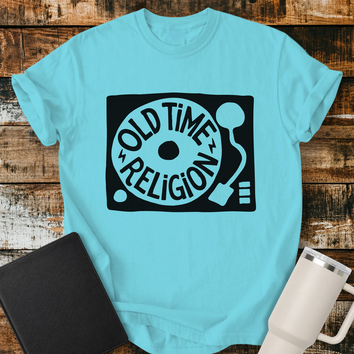 lagoon blue flat-lay t-shirt mockup featuring a bold black-and-white retro record player design with the words “OLD TIME RELIGION” curved around the vinyl record. The vintage-inspired Christian graphic is centered on the shirt, which is displayed on a rustic wood plank background alongside a black leather Bible and a white insulated travel mug.