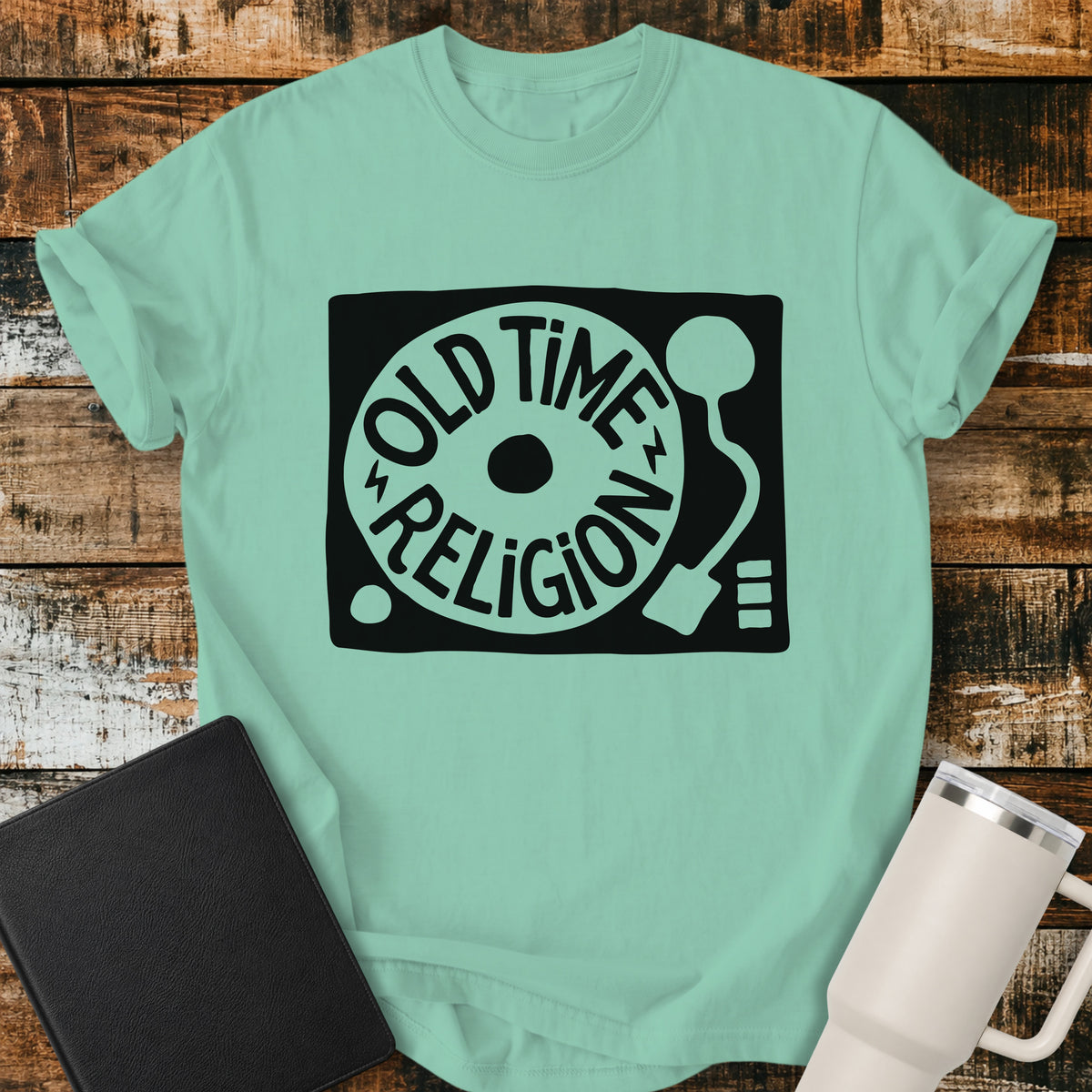 island reef green flat-lay t-shirt mockup featuring a bold black-and-white retro record player design with the words “OLD TIME RELIGION” curved around the vinyl record. The vintage-inspired Christian graphic is centered on the shirt, which is displayed on a rustic wood plank background alongside a black leather Bible and a white insulated travel mug.