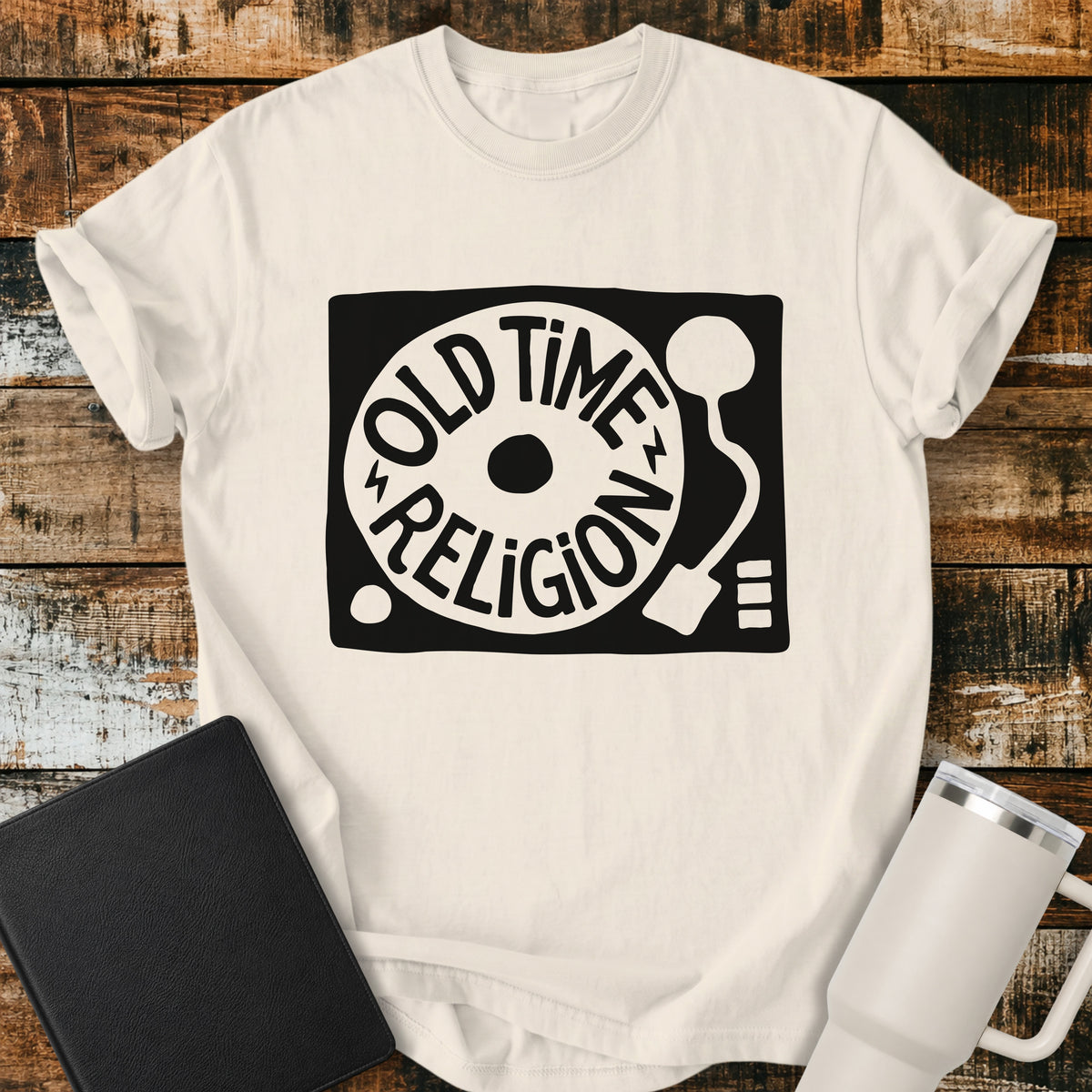 ivory flat-lay t-shirt mockup featuring a bold black-and-white retro record player design with the words “OLD TIME RELIGION” curved around the vinyl record. The vintage-inspired Christian graphic is centered on the shirt, which is displayed on a rustic wood plank background alongside a black leather Bible and a white insulated travel mug.