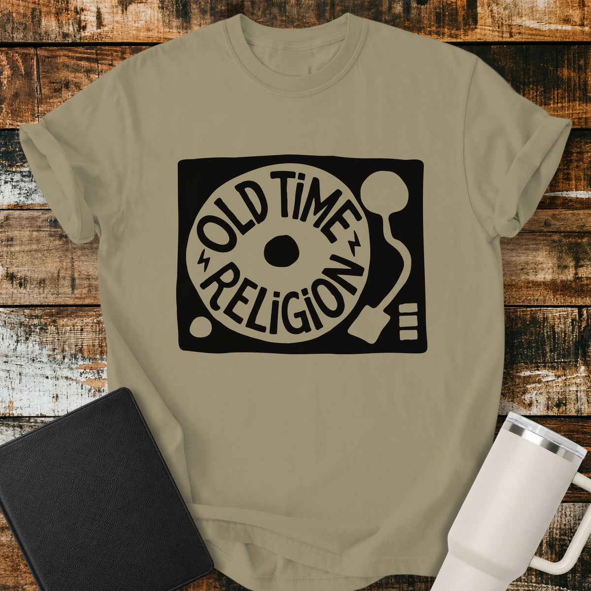 khaki flat-lay t-shirt mockup featuring a bold black-and-white retro record player design with the words “OLD TIME RELIGION” curved around the vinyl record. The vintage-inspired Christian graphic is centered on the shirt, which is displayed on a rustic wood plank background alongside a black leather Bible and a white insulated travel mug.