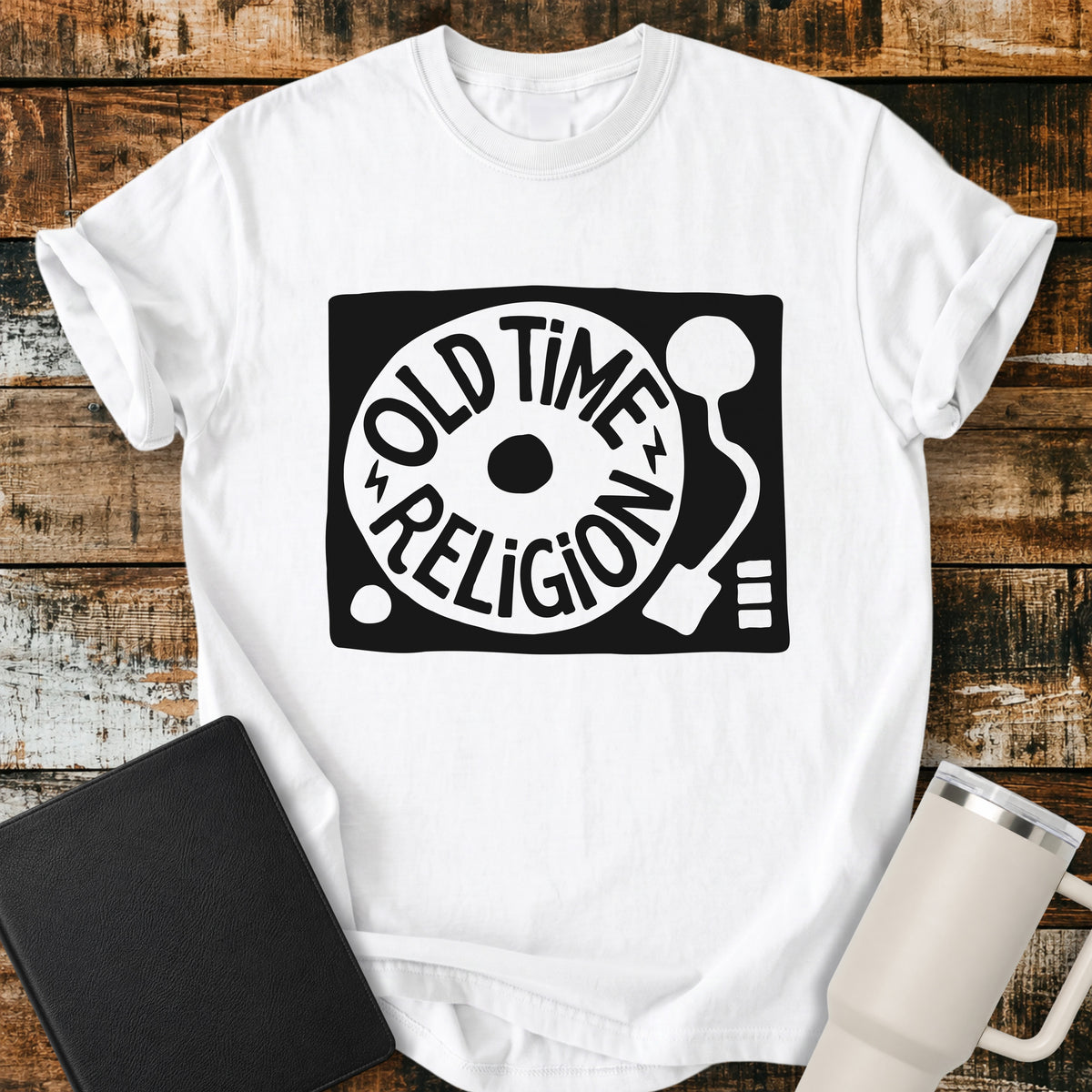 White flat-lay t-shirt mockup featuring a bold black-and-white retro record player design with the words “OLD TIME RELIGION” curved around the vinyl record. The vintage-inspired Christian graphic is centered on the shirt, which is displayed on a rustic wood plank background alongside a black leather Bible and a white insulated travel mug.