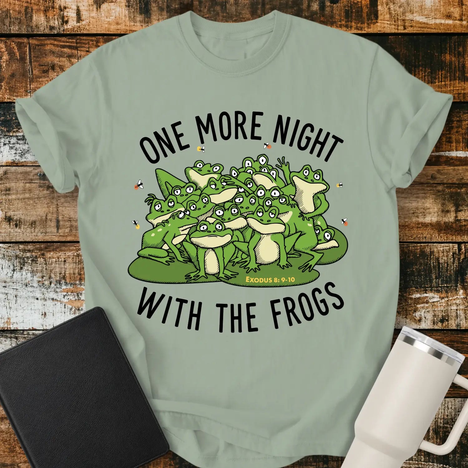 bay green graphic T-shirt mockup featuring a group of cartoon frogs surrounded by fireflies with the phrase “One More Night With the Frogs” and Exodus 8:9–10 text, styled with a black Bible and white tumbler on a rustic wood background.