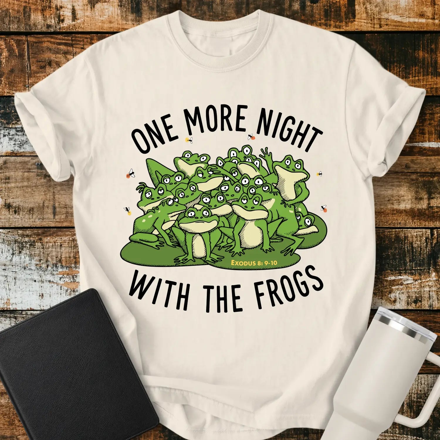ivory graphic T-shirt mockup featuring a group of cartoon frogs surrounded by fireflies with the phrase “One More Night With the Frogs” and Exodus 8:9–10 text, styled with a black Bible and white tumbler on a rustic wood background.