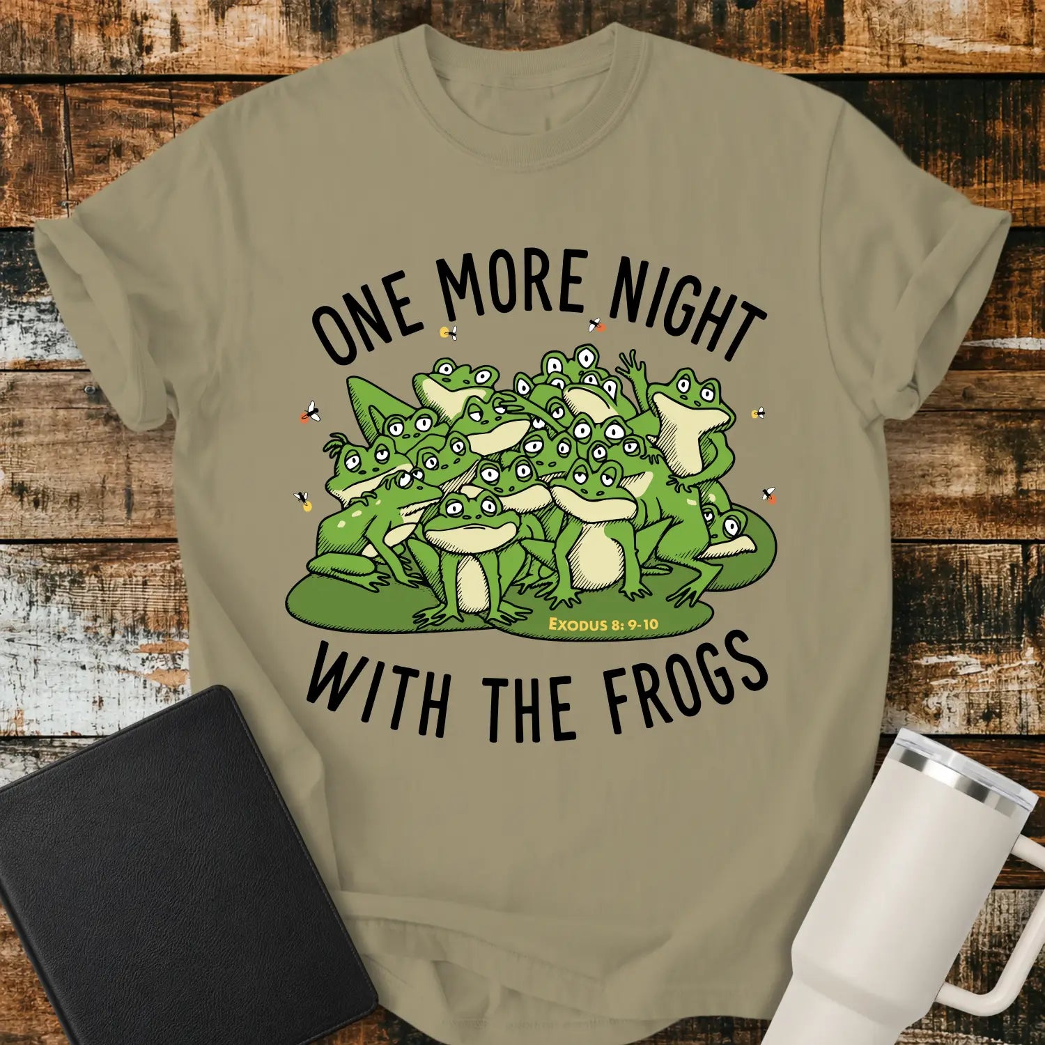 khaki brown graphic T-shirt mockup featuring a group of cartoon frogs surrounded by fireflies with the phrase “One More Night With the Frogs” and Exodus 8:9–10 text, styled with a black Bible and white tumbler on a rustic wood background.