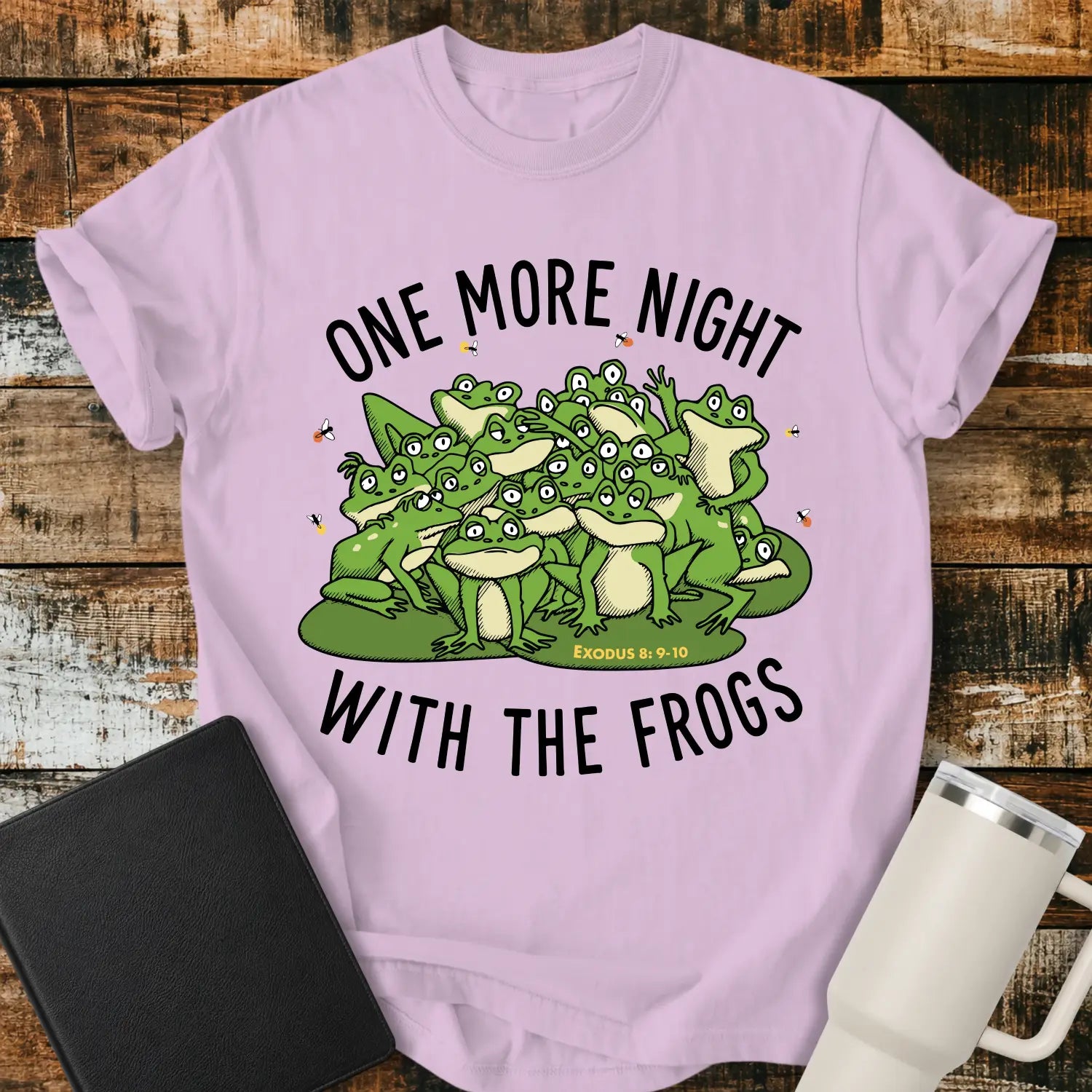 orchid purple graphic T-shirt mockup featuring a group of cartoon frogs surrounded by fireflies with the phrase “One More Night With the Frogs” and Exodus 8:9–10 text, styled with a black Bible and white tumbler on a rustic wood background.