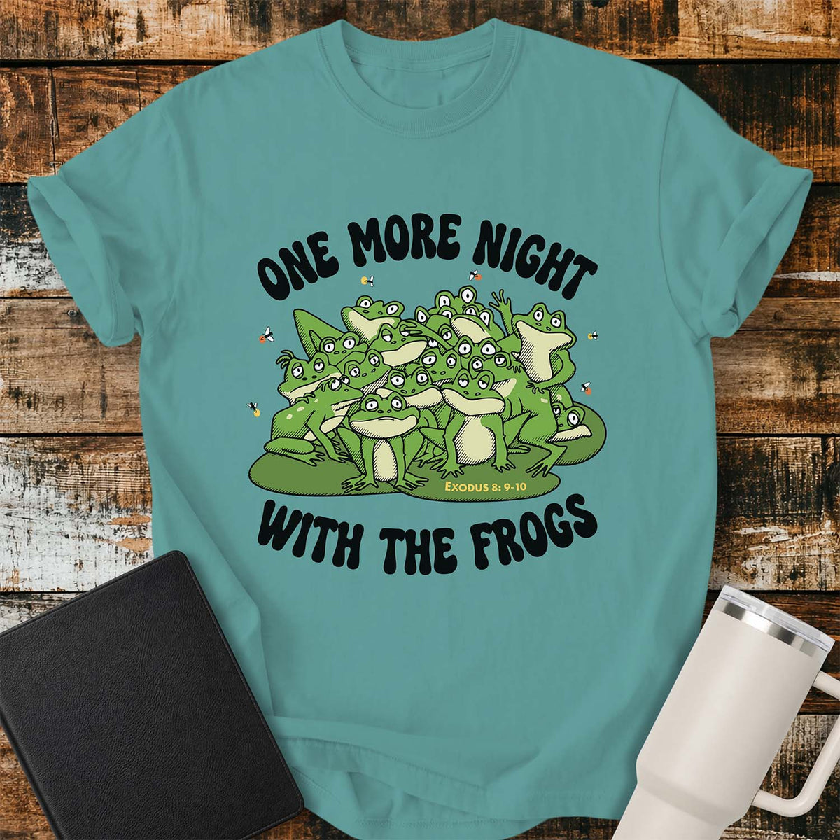 Seafoam green flat-lay t-shirt mockup featuring a playful illustration of a large crowd of cartoon frogs gathered together with small flying insects around them. Bold black lettering reads, "ONE MORE NIGHT" above and "WITH THE FROGS" below, with the Bible reference "Exodus 8:9–10" printed beneath the frogs. The shirt is displayed on a rustic wood plank background alongside a black leather Bible and a white insulated travel mug.