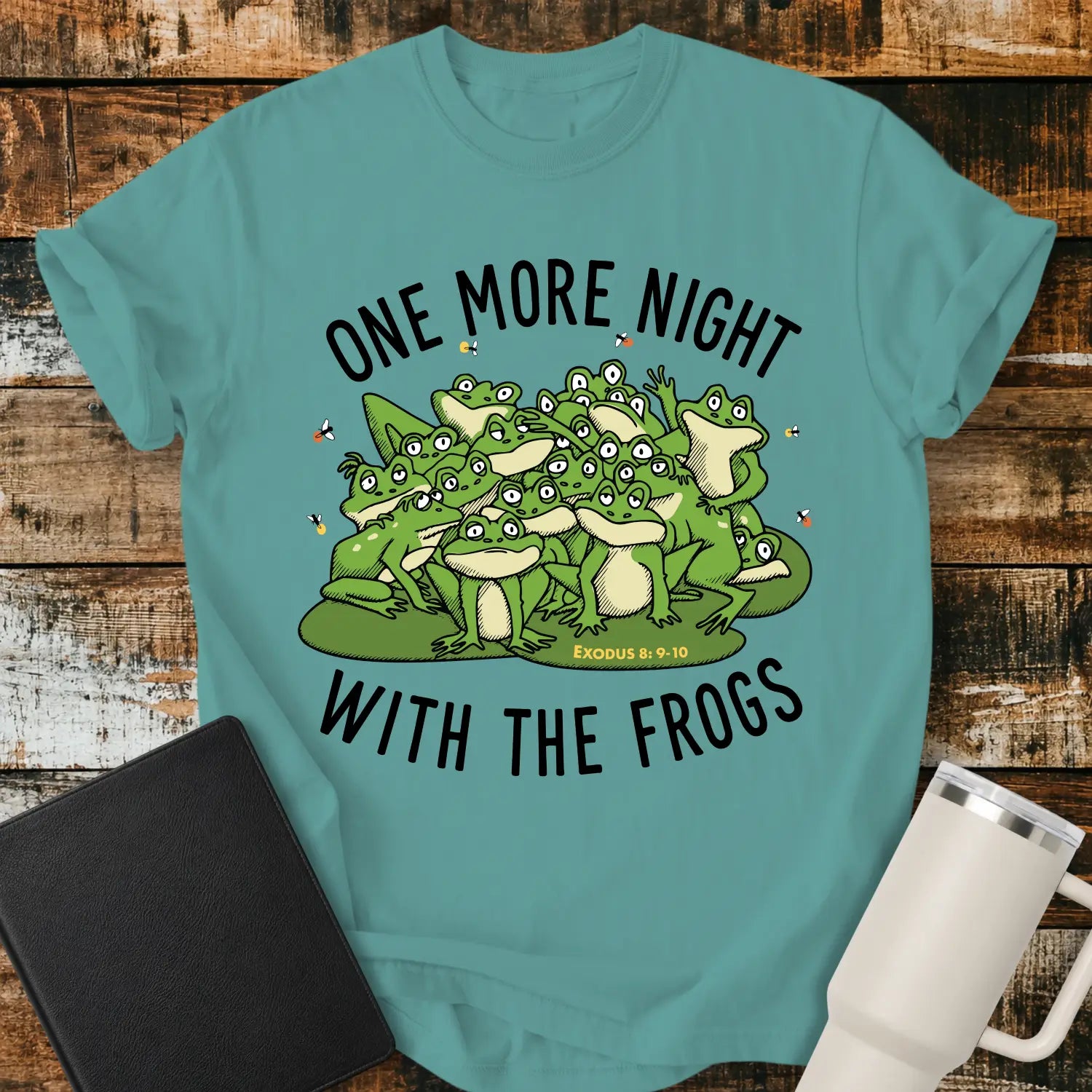 Seafoam green graphic T-shirt mockup featuring a group of cartoon frogs surrounded by fireflies with the phrase “One More Night With the Frogs” and Exodus 8:9–10 text, styled with a black Bible and white tumbler on a rustic wood background.