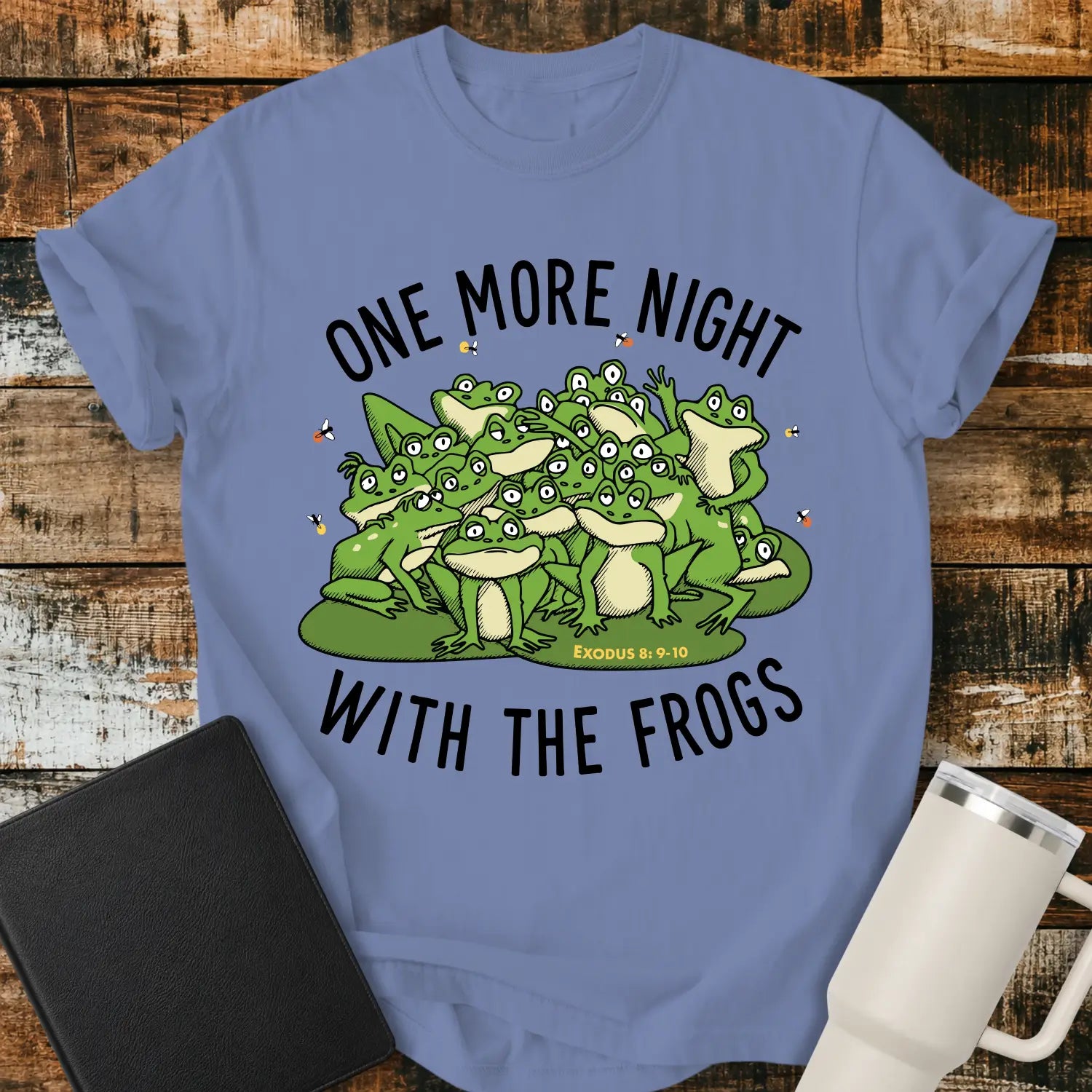 washed denim blue graphic T-shirt mockup featuring a group of cartoon frogs surrounded by fireflies with the phrase “One More Night With the Frogs” and Exodus 8:9–10 text, styled with a black Bible and white tumbler on a rustic wood background.