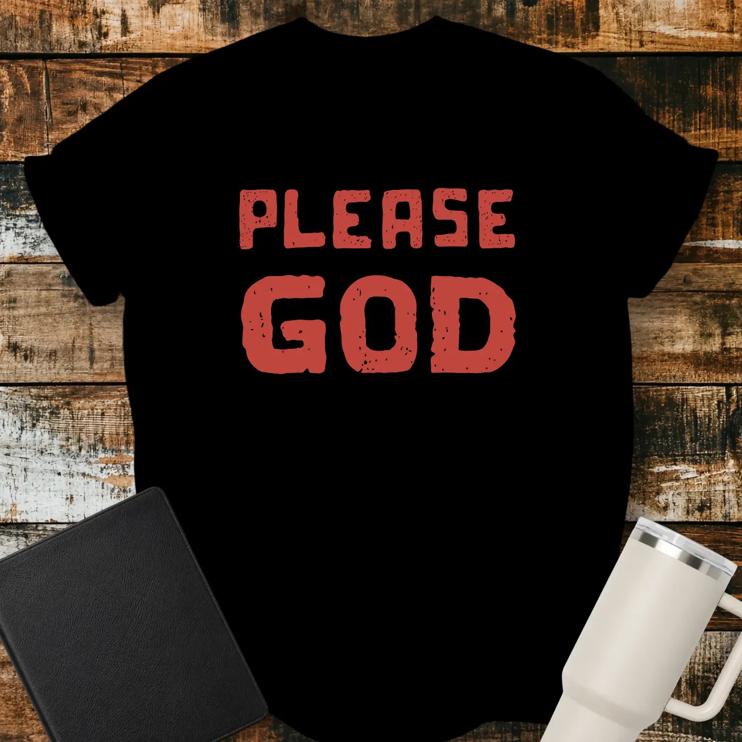 black T-shirt mockup featuring the simple phrase “PLEASE GOD” in large distressed red block lettering. The minimalist design is centered on the shirt, creating a bold faith-inspired statement. The shirt is displayed on a rustic wood plank background beside a black Bible and a white insulated tumbler.