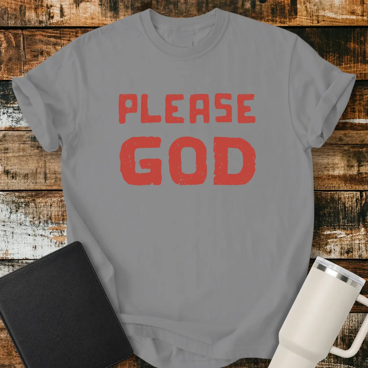 grey T-shirt mockup featuring the simple phrase “PLEASE GOD” in large distressed red block lettering. The minimalist design is centered on the shirt, creating a bold faith-inspired statement. The shirt is displayed on a rustic wood plank background beside a black Bible and a white insulated tumbler.